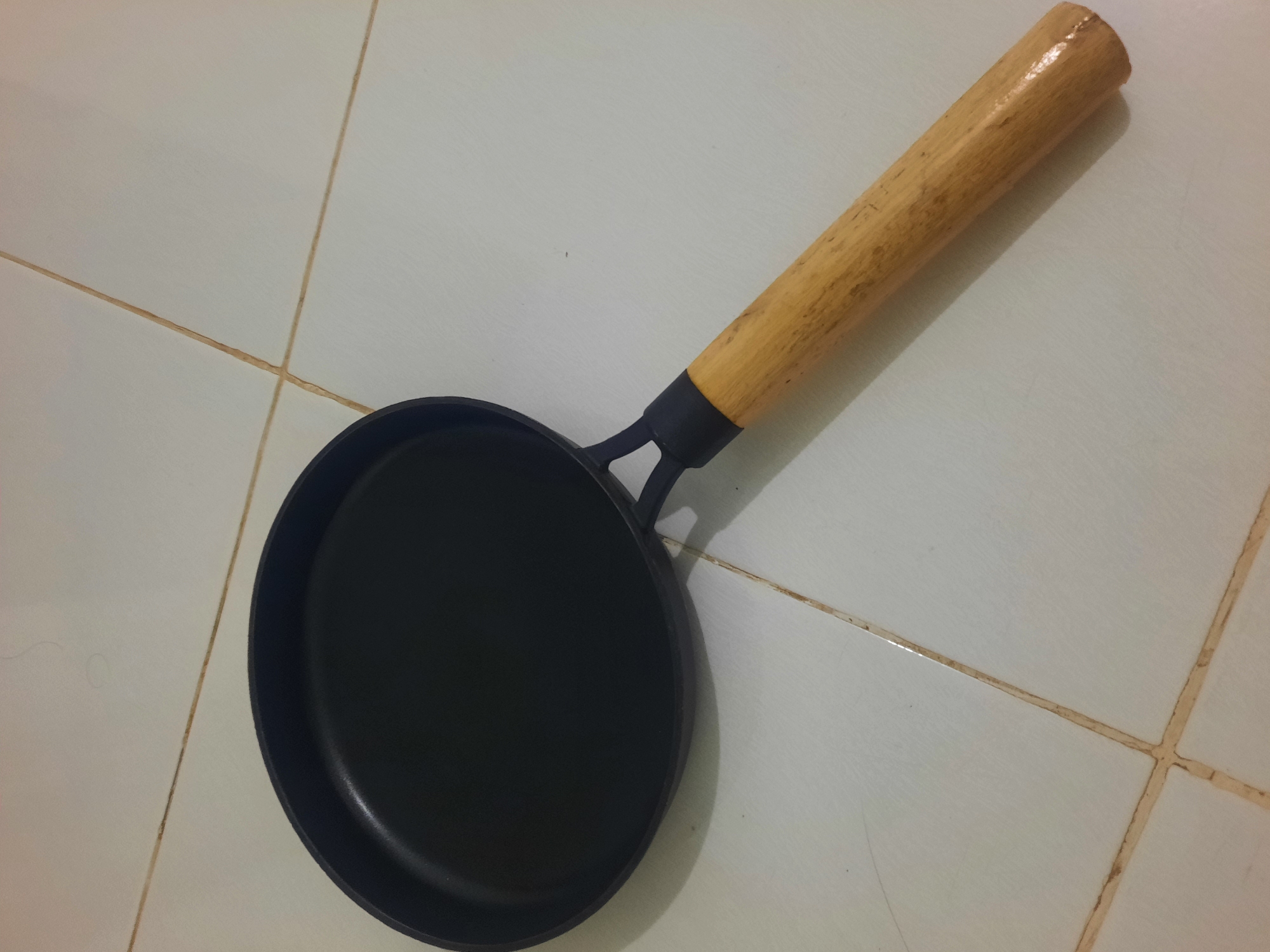 A black frying pan with a wooden handle rests on a tiled floor. The pan is positioned diagonally, with the handle extending towards the bottom right of the image. The tiles are a light color, possibly white or cream, and appear to be square or rectangular. The grout lines between the tiles are visible and are a darker, yellowish-brown color, indicating they may be dirty. There are no people or any other objects in the frame besides the frying pan and the floor. The lighting in the image is somewhat dim, casting subtle shadows. The scene appears to be indoors, likely a kitchen or storage area. The specific location in Lhokseumawe, Indonesia, cannot be determined from the image alone.
