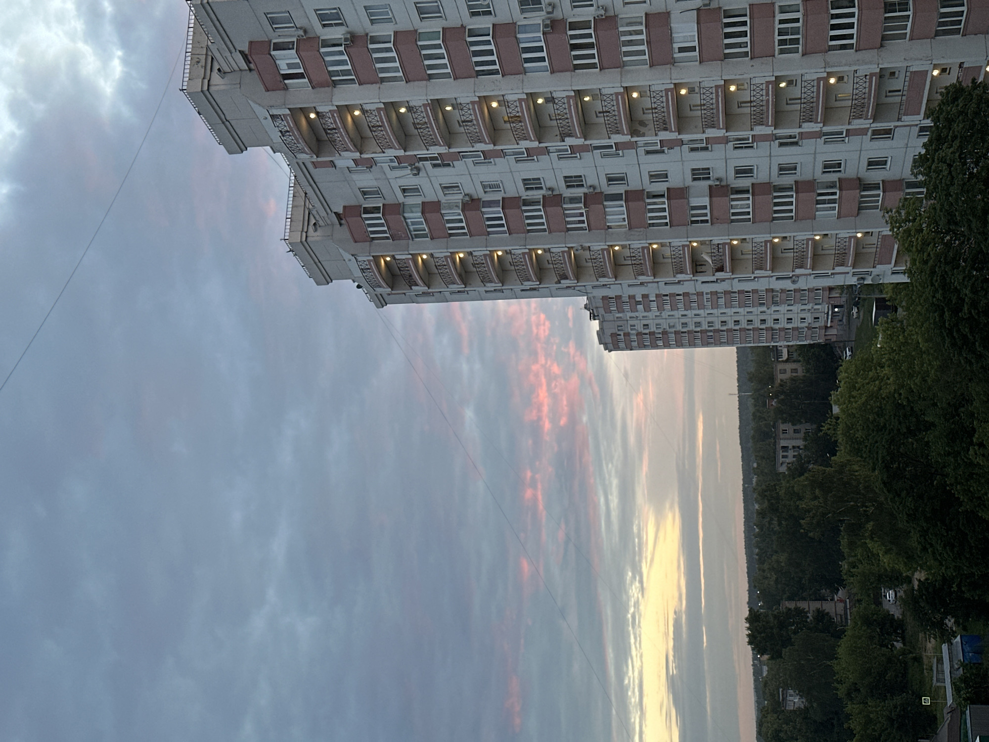 A tall, multi-story apartment building with a light-colored facade and pink accents on the balconies and window frames dominates the right side of the image. The building has numerous windows and balconies with railings. Several lights are on in the windows. To the left of the prominent building, a similar, slightly shorter apartment building is visible, partially obscured.
The background features a dense forest of green trees stretching towards the horizon. In the distance, beyond the trees, several other, lower buildings are scattered. The sky above is filled with clouds. The upper portion of the sky is a muted blue-gray, while the lower portion, closer to the horizon, is illuminated with soft hues of pink and yellow, suggesting either sunrise or sunset. A thin wire stretches diagonally across the sky from the top left to the bottom right of the image.
The scene appears to be a residential area within a city, possibly in the early morning or late evening. The overall impression is one of urban sprawl set against a natural backdrop, with the sky adding a touch of color and atmosphere.