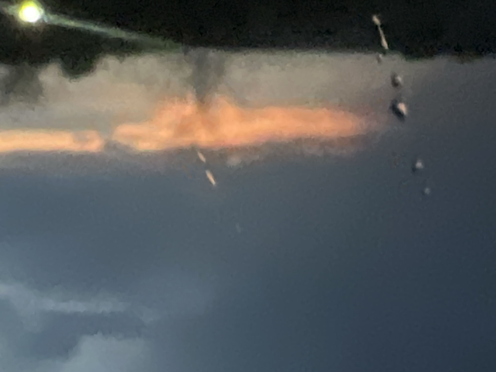 The image shows a blurry night scene, possibly in Votkinsk, Russia. At the top is a dark area with a bright light on the left. Below that, there is an orange-colored atmospheric phenomenon, perhaps clouds illuminated by city lights. The lower part of the image consists of darker and lighter areas, possibly representing clouds or other weather effects. There appear to be a few out-of-focus droplets near the center-right.