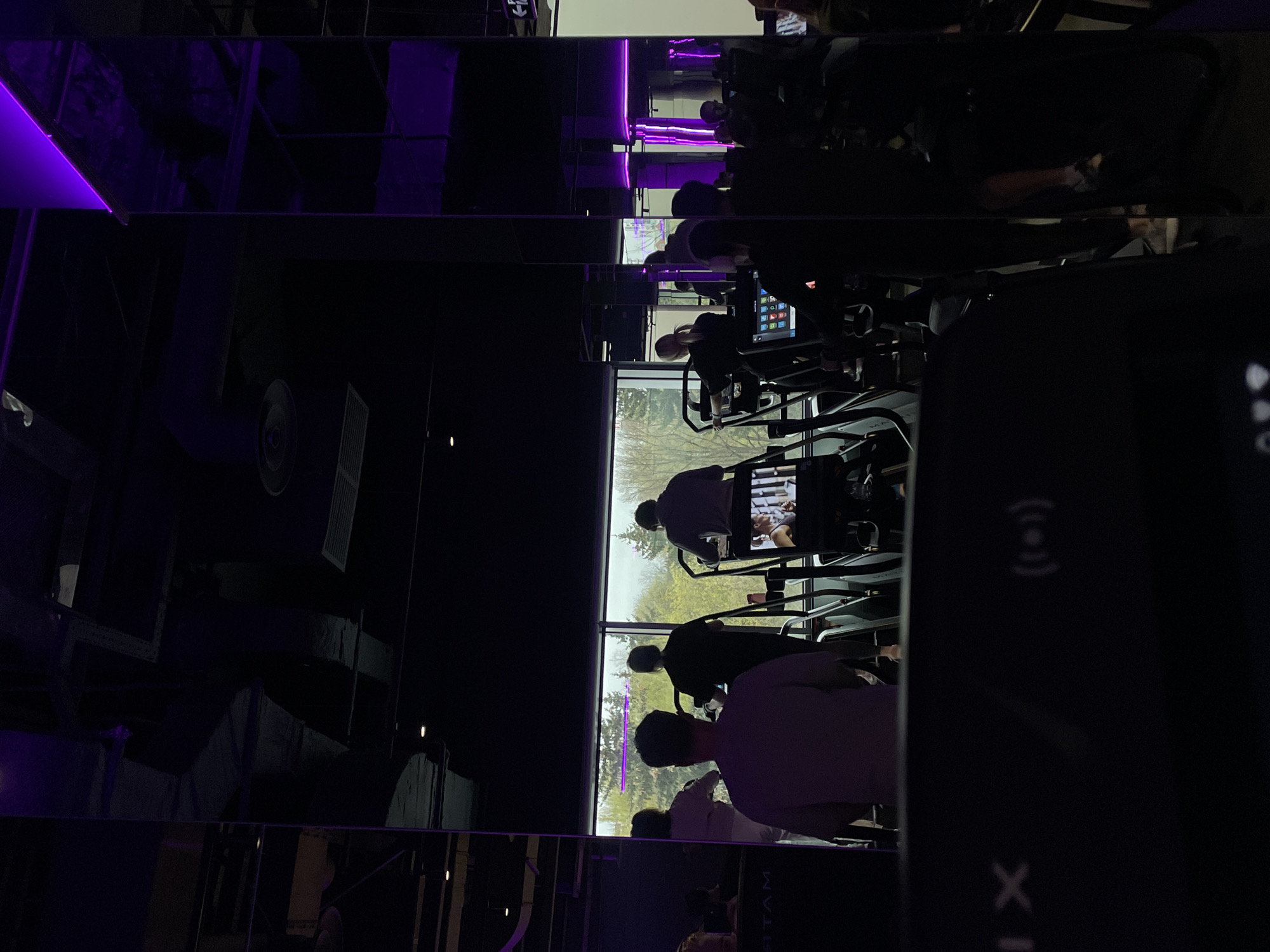 This vertically oriented image depicts the interior of a modern fitness center or gym, likely located in Moscow, Russia. The scene is characterized by dark tones, vibrant purple LED strip lighting, and prominent reflections from what appear to be mirrored or highly polished surfaces on the ceiling or walls.
Several individuals are visible, mostly from behind or the side, engaged in exercising on cardio machines such as treadmills or stair climbers. These machines are arranged in rows facing a large wall of windows, through which a bright, possibly overcast, daytime sky and green trees are visible, suggesting an outdoor natural environment.
The indoor setting features a sleek design with purple LED lighting strips casting a striking glow. Industrial-style elements like air ducts and vents are also discernible on the ceiling. The strong reflections create horizontal bands across the image, distorting and mirroring elements of the room. In the immediate foreground on the right, a portion of a dark smartphone or similar device is visible, showing a Wi-Fi-like symbol and an 'X' icon. The screens on the exercise machines display faint outlines of application icons. The overall ambiance is modern and focused on physical activity.