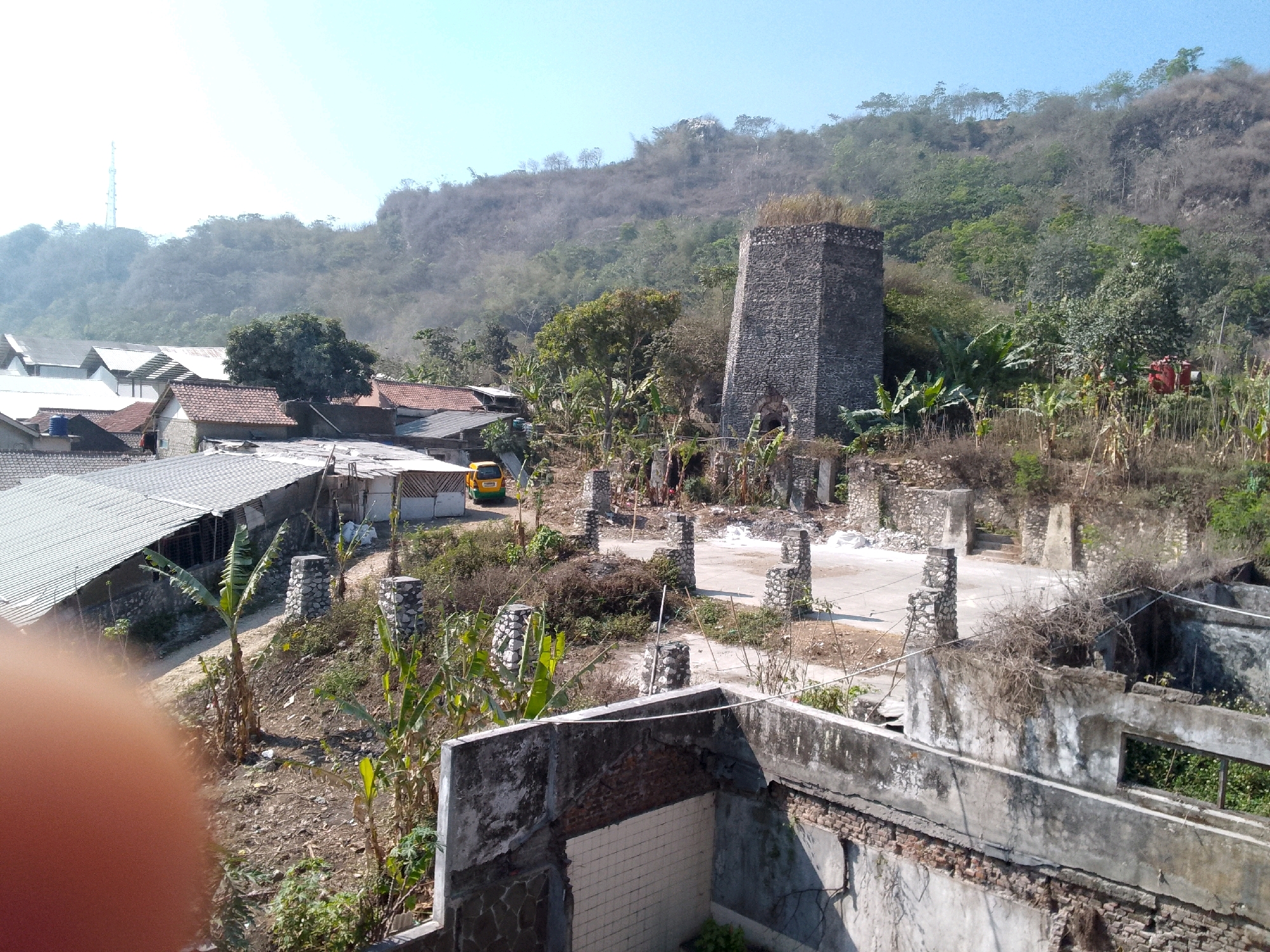 The image depicts a rural or semi-rural landscape in Gunungmasigit, Indonesia. The scene is dominated by a tall, rough-hewn stone structure, possibly an old kiln or furnace, situated on a hillside. In the foreground, there are remnants of weathered concrete structures, suggesting abandonment or disrepair. The middle ground features a collection of houses with tiled roofs and a yellow vehicle. The background consists of a steep, vegetation-covered hillside under a clear blue sky. The environment is characterized by dry earth, scattered trees, and some leafy plants, indicating a possibly arid or dry season. The overall impression is one of a historic or decaying industrial site integrated into a residential area.