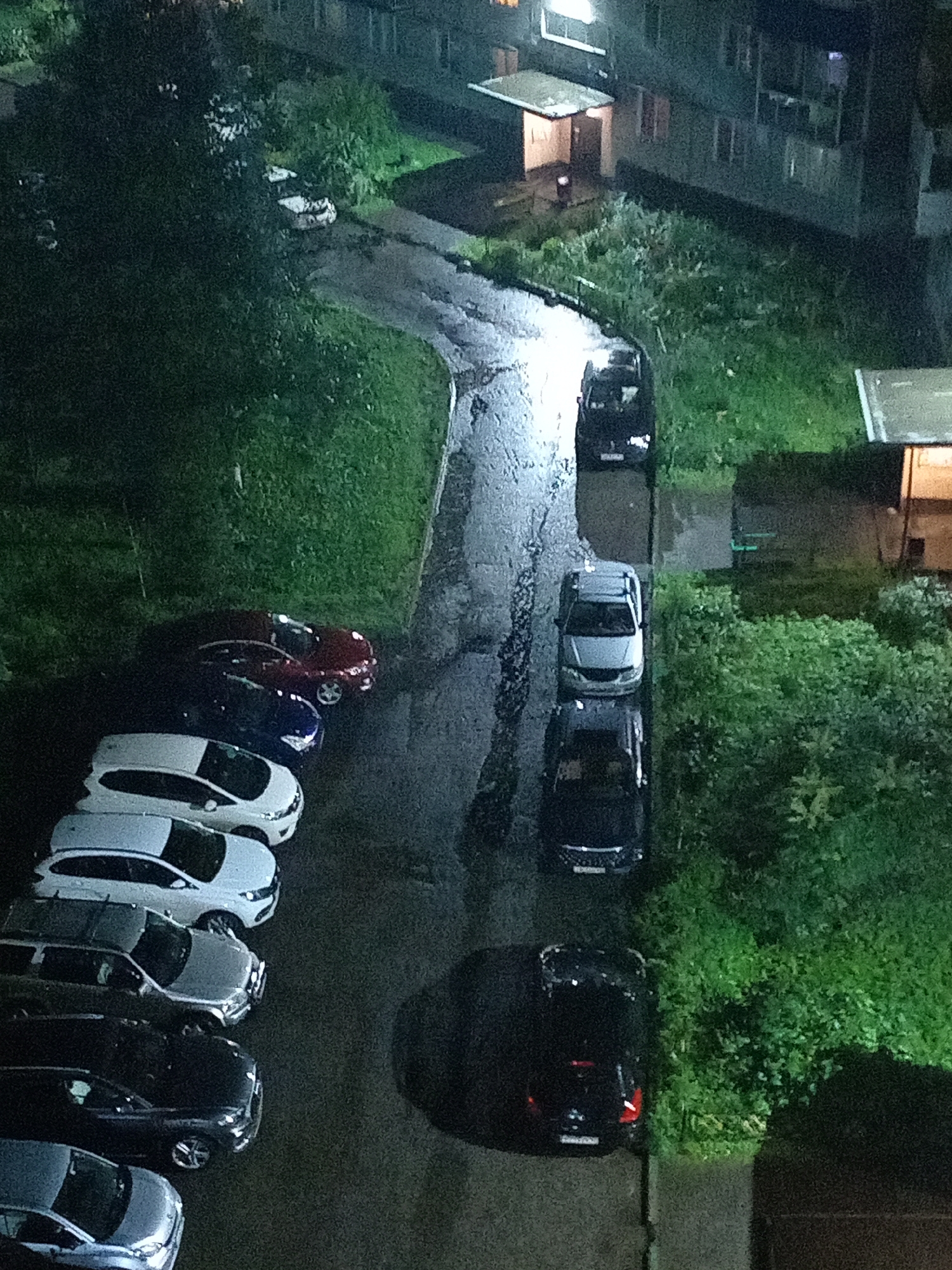 An aerial view shows a dimly lit, wet, asphalt parking lot next to a grassy area and a multi-story building. Several cars are parked in rows, some of which are wet and reflecting the ambient light. In the upper left corner, a white car is partially visible under a tree. Further down, a dark red car and a dark blue car are parked side by side. Below them, a line of white, silver, and black cars are parked in a row. In the upper right of the frame, a dark-colored car is parked in the middle of the paved area. The building in the background appears to be residential, with some windows illuminated. The overall scene suggests a residential area at night, possibly during or after rainfall.