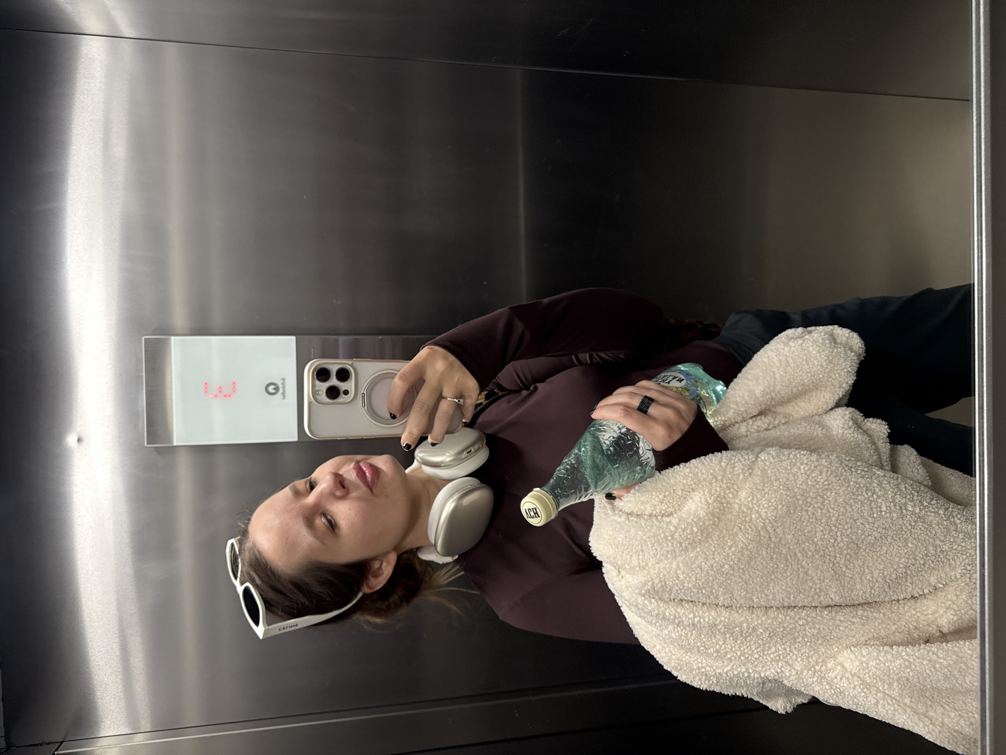 A woman in sunglasses, a maroon top, and headphones poses in an elevator, holding a water bottle and a white, fuzzy jacket. The elevator panel displays red numbers, and the surrounding surfaces are stainless steel. The location cannot be determined to be Barcelona, Spain, from the image alone.