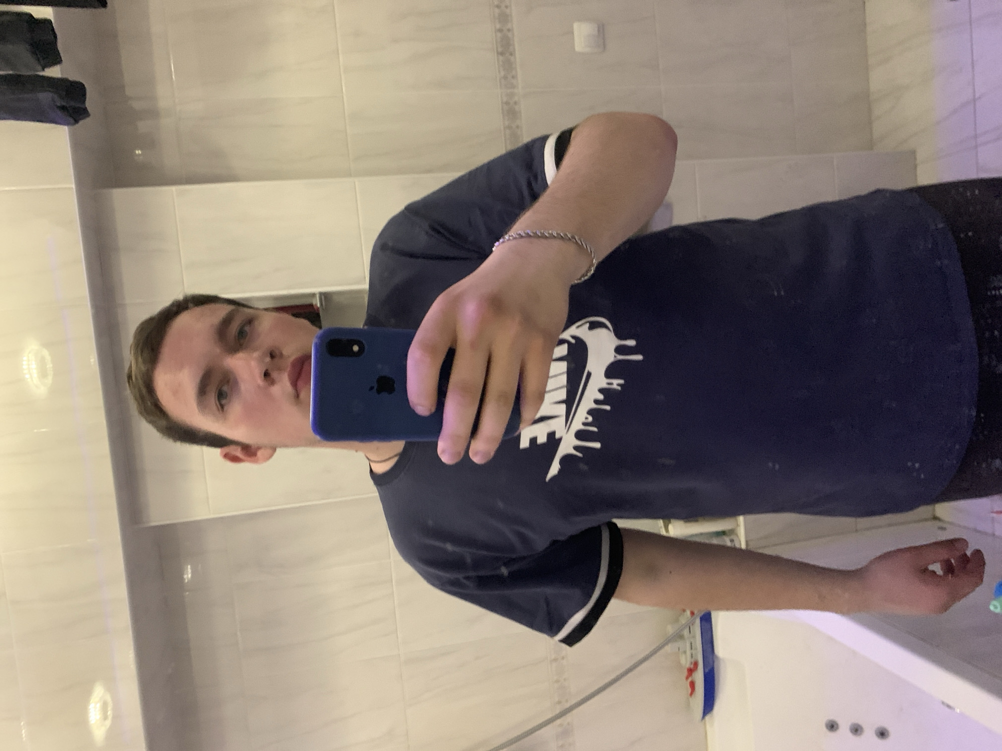 A young man stands in a bathroom taking a mirror selfie with a blue smartphone. He wears a dark blue t-shirt with a white Nike logo and white-striped sleeves. A silver chain bracelet is on his left wrist. The bathroom has white tile walls. A white bathtub is partially visible in the bottom right corner. The image does not provide evidence of Severodvinsk, Russia.
