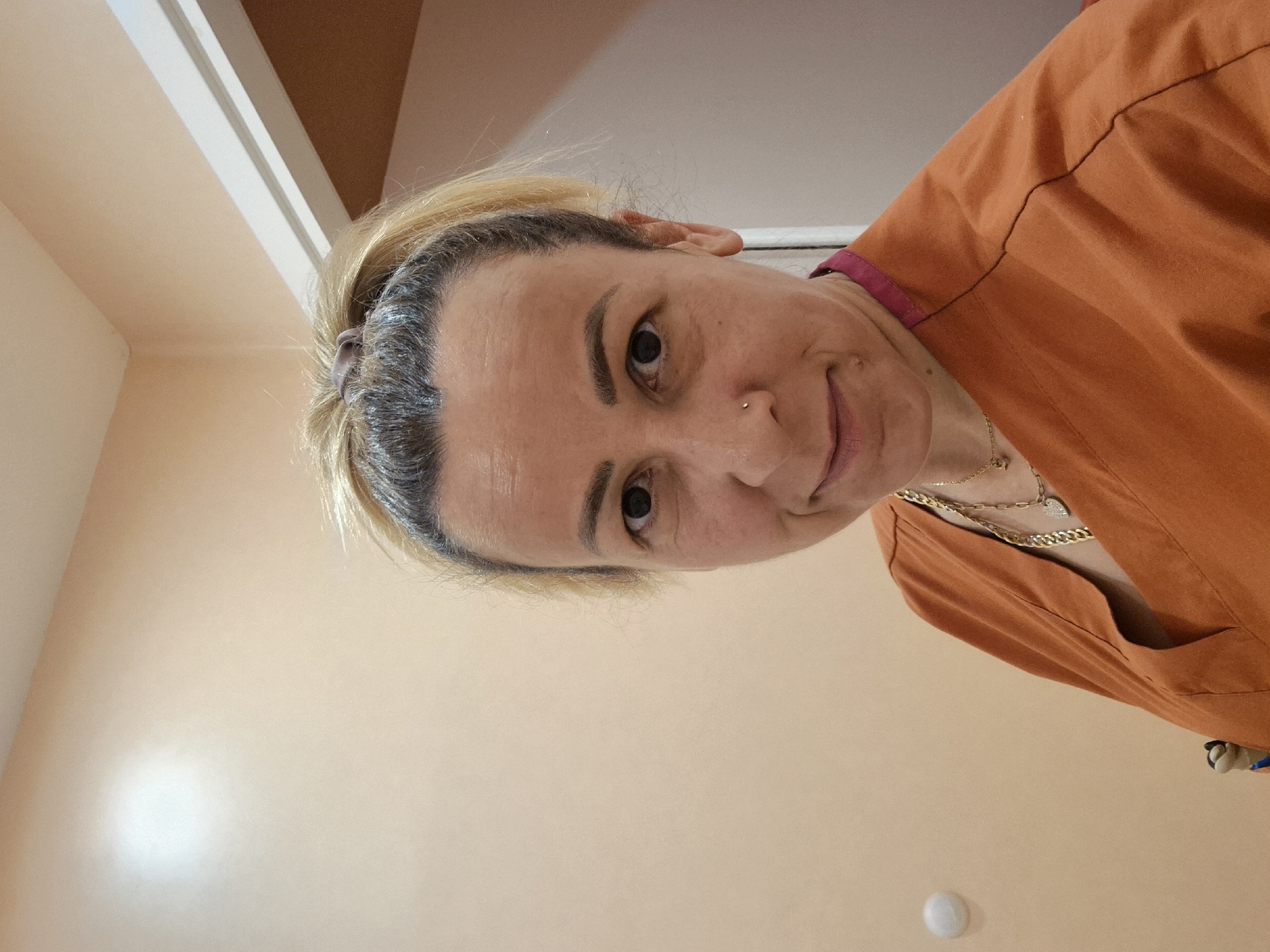 An adult female is depicted in a close-up, selfie-style indoor portrait in the city of Unknown, Unknown. She has blonde hair pulled back into a high ponytail, revealing darker roots, and a direct gaze towards the camera with a subtle, closed-mouth smile. Her face exhibits visible wrinkles around the eyes and forehead, and she has a small silver stud piercing on her left nostril. She is wearing a burnt orange V-neck uniform top, possibly medical scrubs, with a fuchsia trim visible along the inner collar. Multiple gold necklaces are worn around her neck, including a thick curb chain, a thinner chain with a heart pendant, and another thin chain with a small round charm. The background consists of plain light peach-colored walls. A prominent circular bright light reflection is visible on the upper left wall. A partial white door frame or trim is visible on the right side of the image, and a small, round, white object is affixed to the lower left wall.