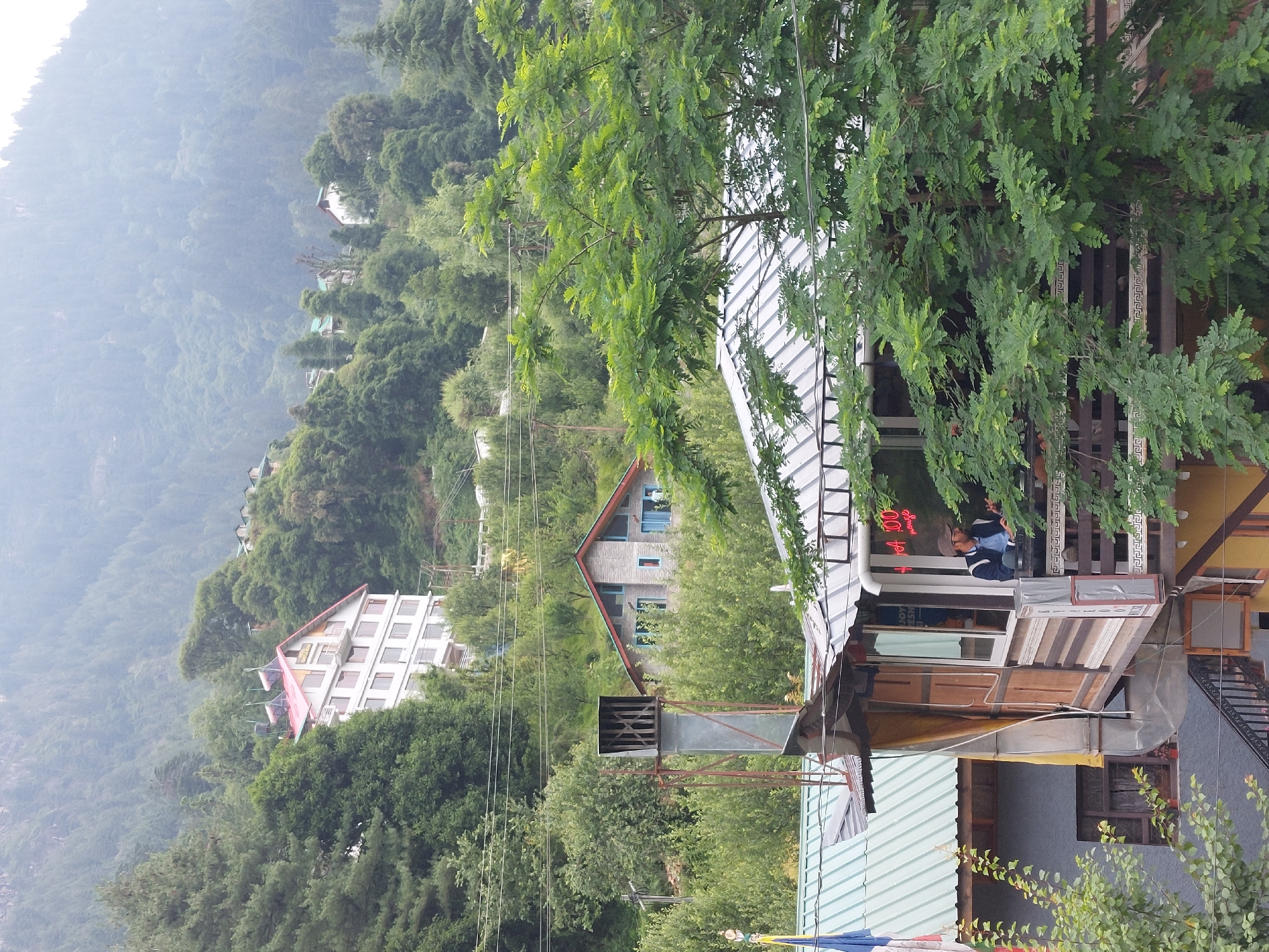 The image captures a view of the town of Vashisht, India, nestled amidst lush green hills and dense forests. In the foreground, a multi-story building with a distinctive red roof is partially obscured by a large tree. This building appears to be a hotel or guesthouse, with solar panels visible on its roof. Below it, another structure with a corrugated metal roof is visible, featuring a prominent metal chimney stack. A person is seen standing on the balcony of this lower building, looking out. Further in the background, another building with a light-colored facade and a red roof is perched on the hillside, surrounded by trees. The overall scene depicts a mountainous, verdant landscape with a scattering of buildings, suggesting a popular tourist destination. The atmosphere appears to be somewhat hazy, possibly due to mist or smog.