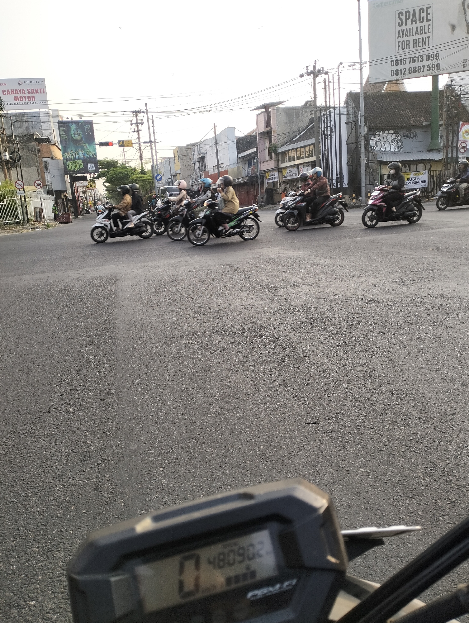 The image captures a street scene in Surakarta, Indonesia, focusing on an intersection with moderate traffic. Several motorcycles with riders are present, some waiting at what appears to be a traffic light. The foreground shows a portion of a motorcycle's dashboard, displaying the numbers "0" and "480902". Buildings line the street, with various signs visible, including advertisements and rental notices. The sky is overcast, suggesting it is either early morning or late afternoon. The road surface is asphalt, and power lines are strung overhead. The overall atmosphere is one of everyday urban activity.