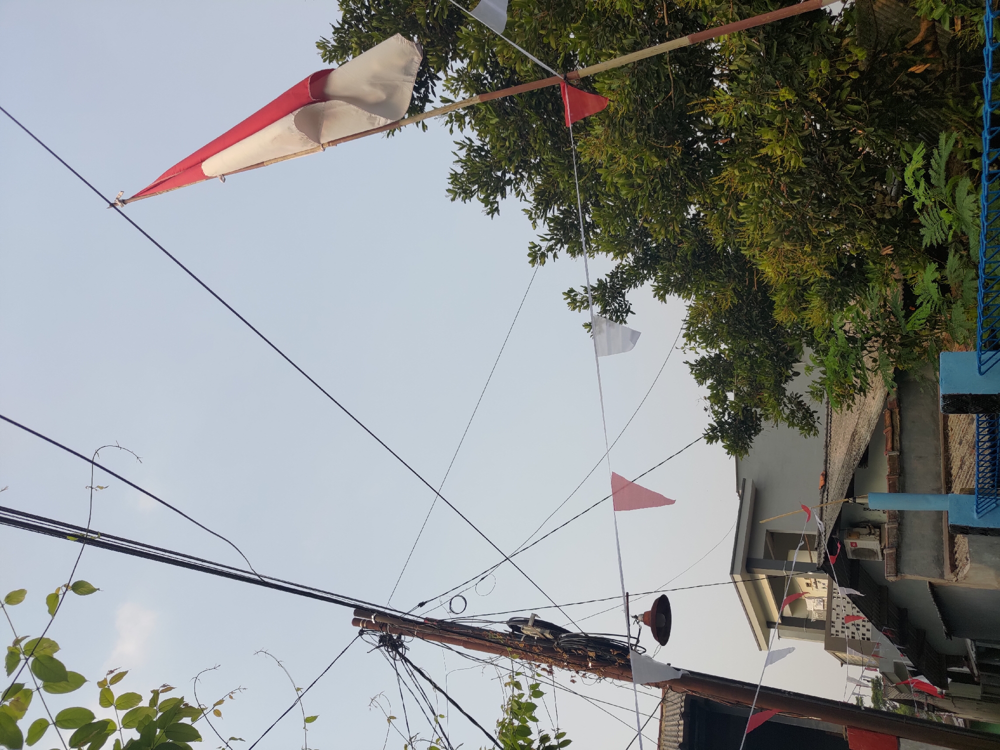 In Depok, Indonesia, a flag of Indonesia flies from a bamboo pole against a pale sky, its red and white colors vibrant despite the overcast conditions. The flag is positioned to the right of a utility pole that is laden with a dense network of wires, suggesting the presence of electricity and telecommunications infrastructure. A vintage-style lamp hangs from the utility pole, casting a shadow on the ground below.
Along a wire strung across the scene, triangular flags in red and white, reminiscent of celebratory decorations, are displayed. These flags are arranged in a repeating pattern, adding a festive atmosphere to the urban environment. In the background, a multi-story building with a gray facade and a flat roof is visible. The building's architecture suggests a residential or commercial structure typical of the area. Lush green trees with dense foliage are interspersed throughout the scene, providing a natural contrast to the man-made elements. The ground level is partially obscured by foliage and a blue metal fence, hinting at a residential area or public space.