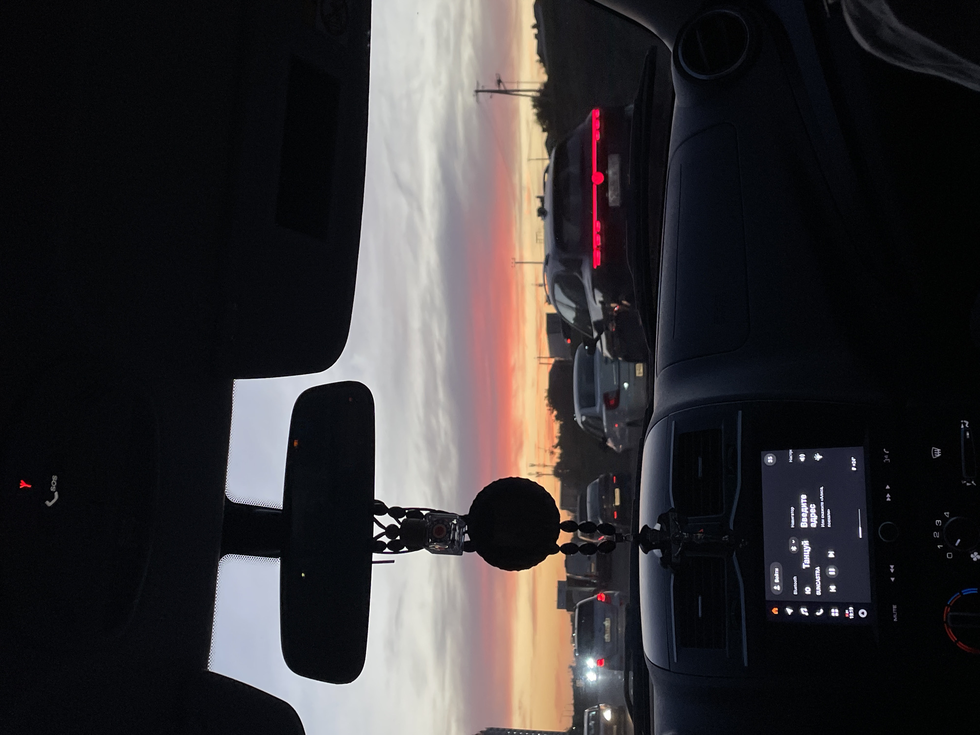 The image, taken from inside a vehicle, captures a traffic jam on a city street in Rostov-on-Don, Russia, during a sunset. The sky is a vibrant mix of orange, pink, and gray, with clouds streaking across. Multiple cars are visible on the road, some with their headlights on. A car in the foreground has a distinctive taillight design. Inside the car, the dashboard displays a navigation screen with a map and music controls. A small cross ornament hangs from the rearview mirror. The overall scene depicts a common urban scenario of a commute in congested traffic at dusk.