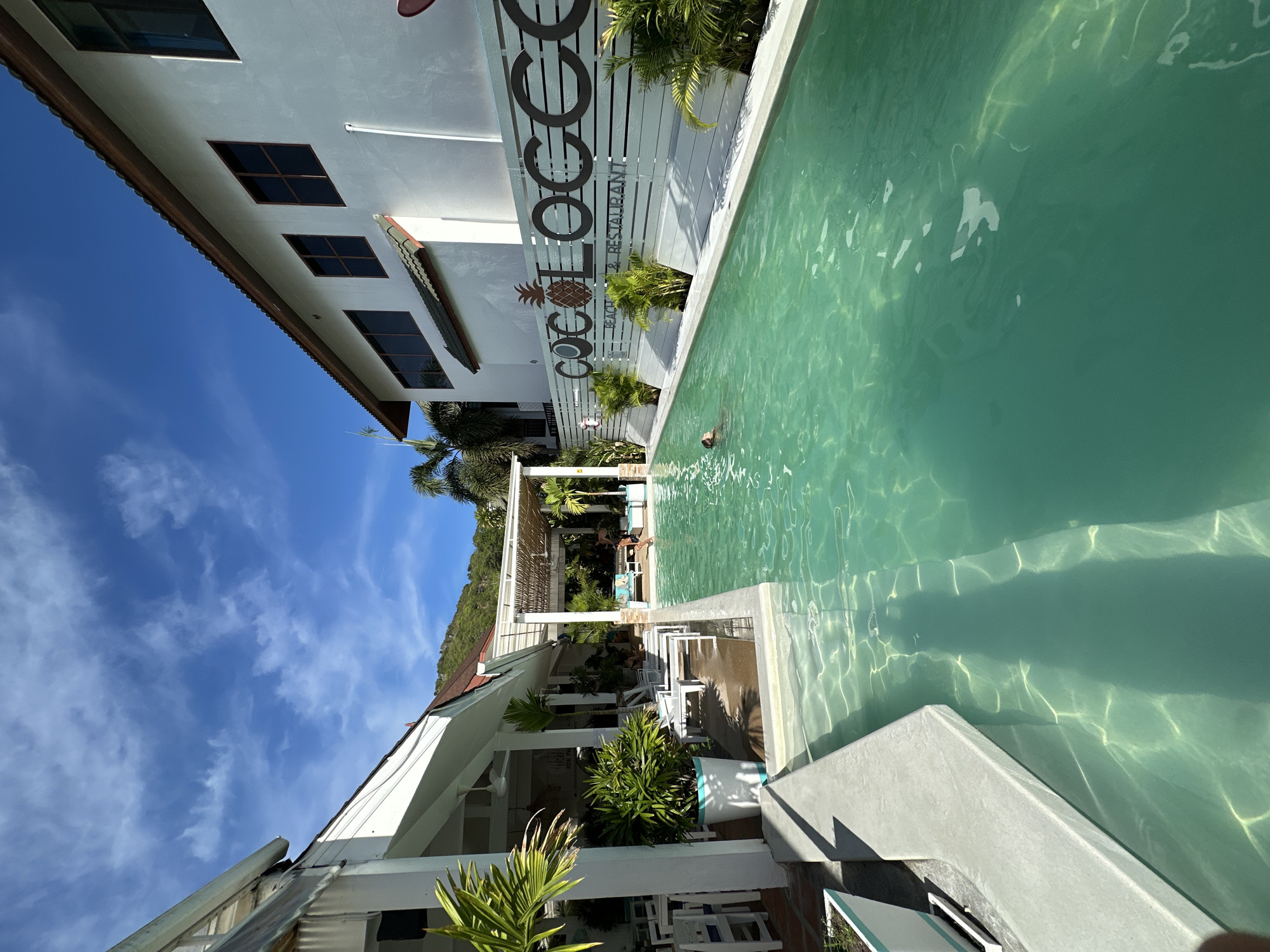 This media file shows a resort with a swimming pool and buildings. The scene is outdoors during the day, with clear blue skies and scattered clouds, indicating fair weather. The location appears to be a tropical setting, as evidenced by the presence of palm trees and lush greenery.
In the foreground, a swimming pool with turquoise water takes up a significant portion of the frame. Sunlight creates patterns of light and shadow on the water's surface. A person is partially visible in the water on the far side of the pool. Adjacent to the pool is a building with white walls and dark-framed windows. A fence with text and a pineapple logo is also visible. The text on the fence reads "COCO LOCO BEACH & RESTAURANT".
Beyond the pool area, there are more buildings with white walls and pitched roofs, some with open-air structures and outdoor seating. There are plants and furniture arranged in these areas. In the background, a lush green hillside is visible, characteristic of a tropical island. The overall atmosphere suggests a relaxing vacation or resort environment. The image is taken from a low angle, looking upwards towards the sky and across the pool area.