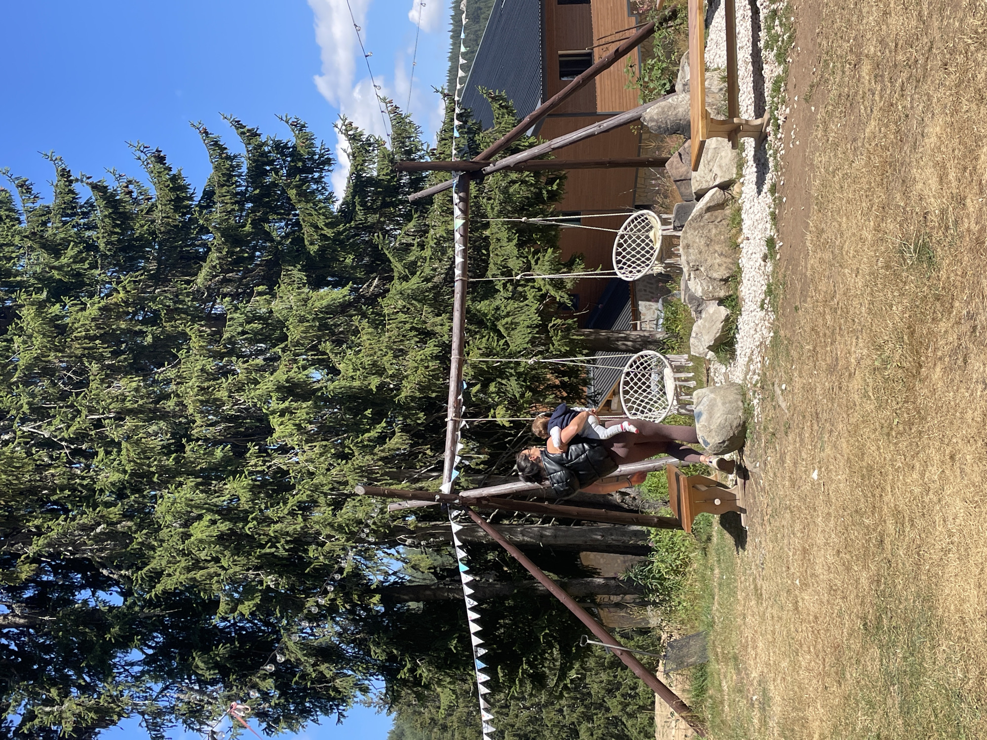A woman carrying a child stands near a wooden swing set in a grassy area. She is wearing a dark jacket and leggings. The swing set has two white woven chairs hanging from it. In the background, there are large pine trees, a wooden building with a dark roof, and a clear blue sky with some clouds. A string of small lights is strung between the trees. The ground is covered in dry grass and scattered rocks. This scene appears to be set in an outdoor recreational area, possibly a park or a resort, located in or near the city of Smolyan, Bulgaria.
