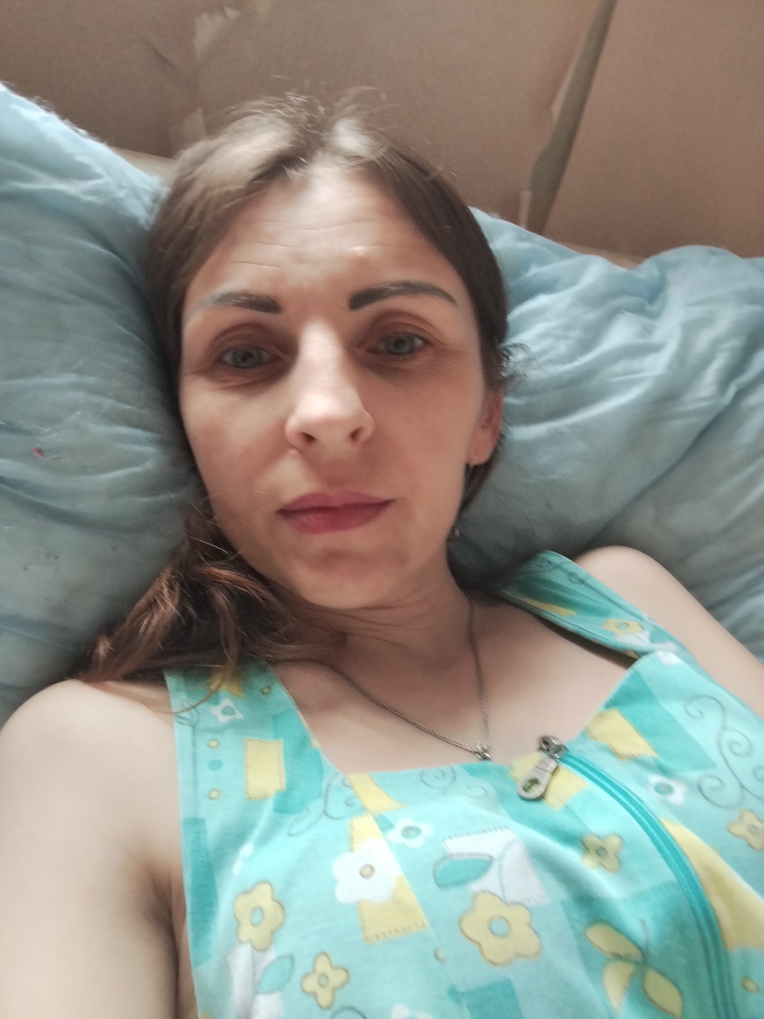 A woman with brown hair and fair skin, wearing a light blue and yellow floral patterned garment with a zipper, is lying in a bed. She is looking directly at the camera. The background consists of pillows and what appears to be a wall or headboard. The scene is indoors, likely in a bedroom.