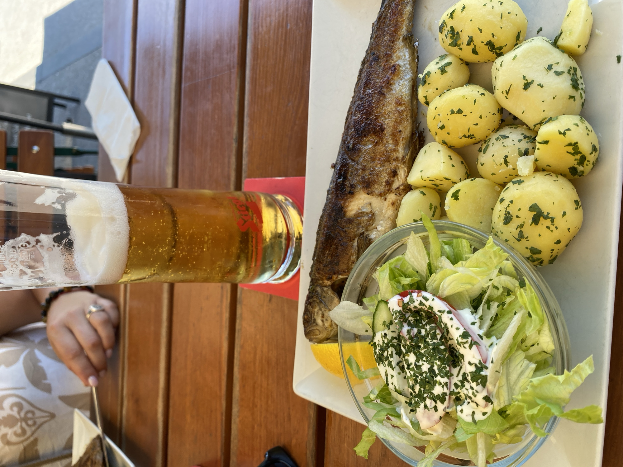 A close-up, eye-level shot shows a meal of whole grilled fish, small boiled potatoes, and a side salad on a white plate, placed on a wooden table. A glass of beer with a frothy head is positioned behind the plate. A person's hand, adorned with a ring, is visible in the background, holding a fork over another plate, implying a shared meal. The meal is presented outdoors, likely at a restaurant in Hallstatt, Austria, given the traditional setting suggested by the wooden furniture and glassware. The overall scene captures a casual dining experience on a sunny day.