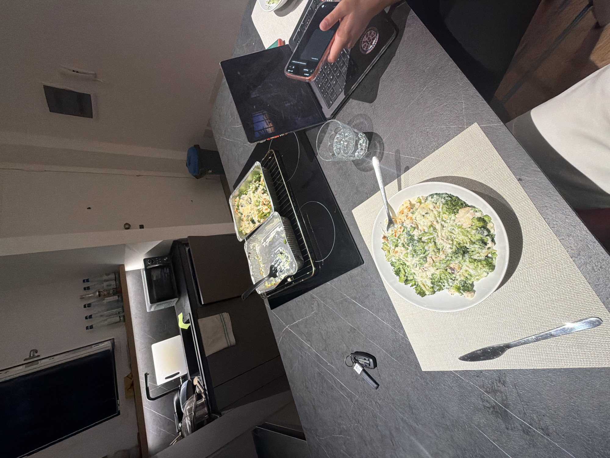 An overhead, medium shot captures a kitchen counter in Milan, Italy, illuminated by ambient light. A person's hands are visible in the foreground, holding a mobile phone and interacting with a laptop. Next to the laptop sits a glass of water and a plate of food. A serving dish containing food is placed on the induction stovetop. In the background, various kitchen appliances and utensils are visible. A set of keys rests on the counter near the food. The overall scene depicts a domestic setting, likely a kitchen or dining area, with evidence of a meal being prepared or consumed alongside digital device usage.