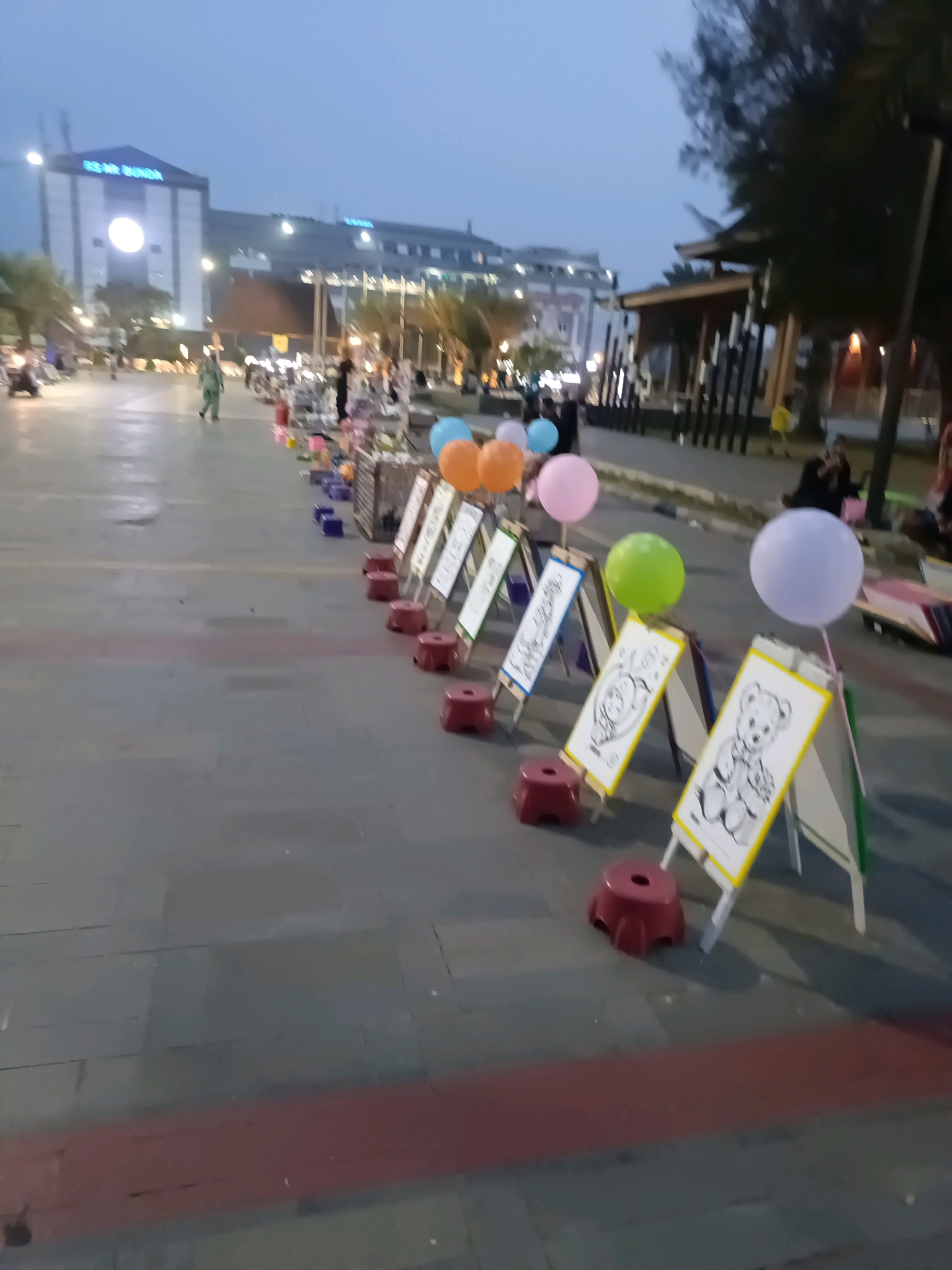 The image depicts a pedestrian walkway in front of a building identified by the sign "RS M. BUNGA," likely indicating a hospital. The walkway appears to be a public space in the city of Lubuklinggau, Indonesia, possibly a park or a promenade given the presence of vendors and recreational elements. The time of day appears to be dusk or early evening, as indicated by the dim lighting and the blue-toned sky.
In the foreground, a row of easels is set up, displaying children's drawings or coloring pages, some featuring cartoon characters like bears and animals. Red stools are placed in front of the easels, suggesting a setup for children to sit and draw. Colorful balloons – pink, orange, light blue, green, and white – are attached to some of the easels and also float freely, adding a festive atmosphere.
Further down the walkway, several vendors appear to be operating stalls, with various items displayed. People are seen walking or browsing, and some are seated. The background reveals a modern building with illuminated signage and a canopy structure with vertical elements, possibly part of a retail or entertainment complex. Trees are visible on the right side of the image, and the ground is paved with concrete slabs, with a distinct red stripe running horizontally across the walkway. The overall scene suggests a lively evening activity in a well-developed urban area.