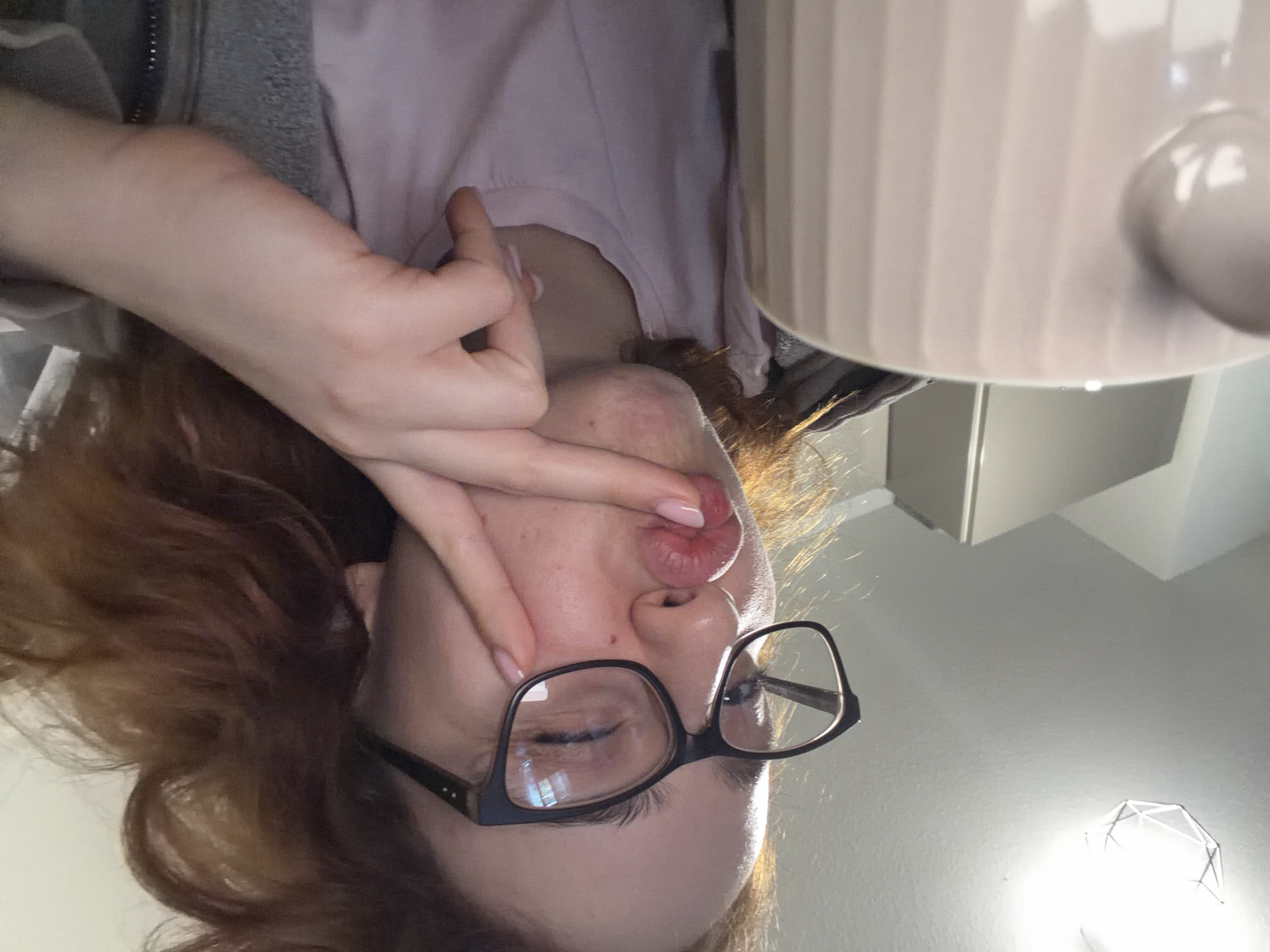 A young woman with auburn hair and glasses is taking a selfie. She is wearing a pink t-shirt and a grey hoodie. Her right hand is on her face, touching her cheek. She is making a kissing face. In the foreground, there is a light pink mug with vertical ridges. The background shows a ceiling with a geometric light fixture and a sliver of a grey kitchen cabinet. The scene appears to be indoors, likely in a residential kitchen. There is no information to suggest this image is located in Strasbourg, France.