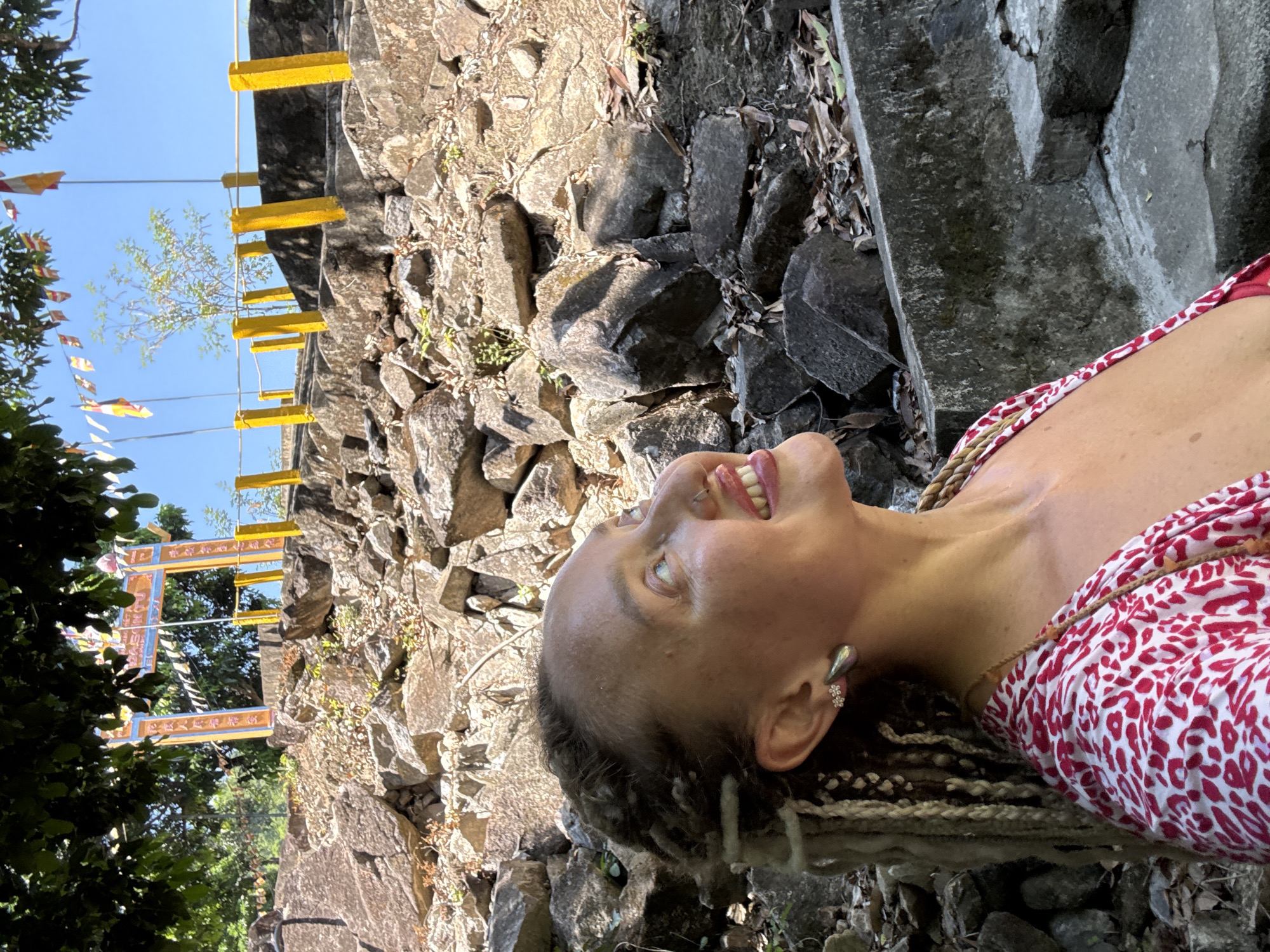 A woman with blonde dreadlocks and a red and white patterned dress is looking up and to the right, with her mouth open in a smile. She is wearing a silver teardrop earring and a nose ring. In the background, there are large rocks and a set of stairs with yellow posts and a railing. Behind the stairs, there is an archway with colorful flags and banners, and more flags on poles. The sky is blue and clear. The scene appears to be at a temple or historical site, possibly in Nha Trang, Vietnam, given the style of the archway and flags which are often seen at Buddhist sites.