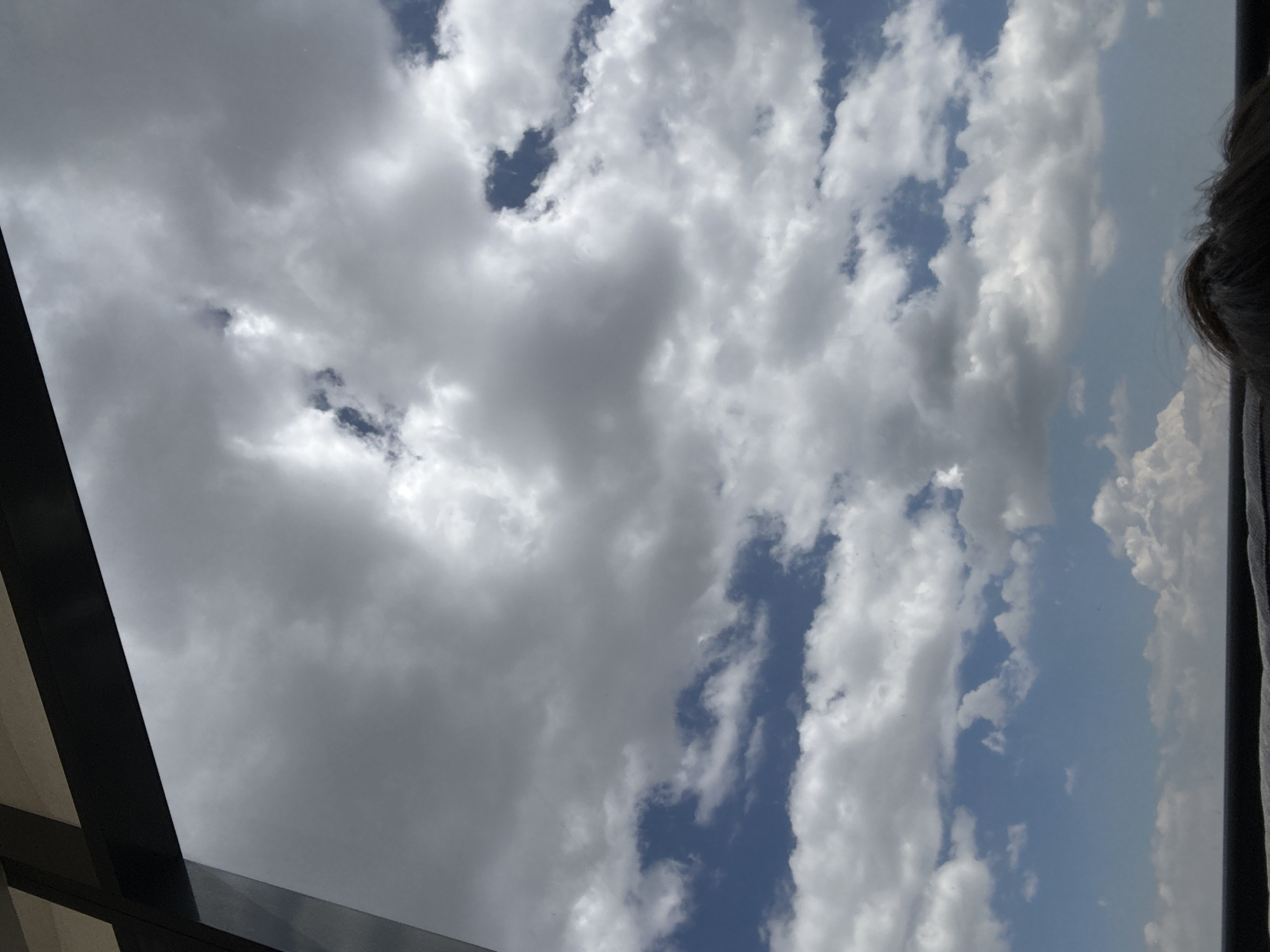 The image shows a view of the sky with clouds, likely taken from inside a building in Huixquilucan de Degollado, Mexico. The sky is partially cloudy, with some areas of blue sky visible between the clouds. Some structural elements, possibly part of a window or roof are visible in the corners of the frame, partially obstructing the view. There is a hint of a human head/hair on the right-hand side of the image.