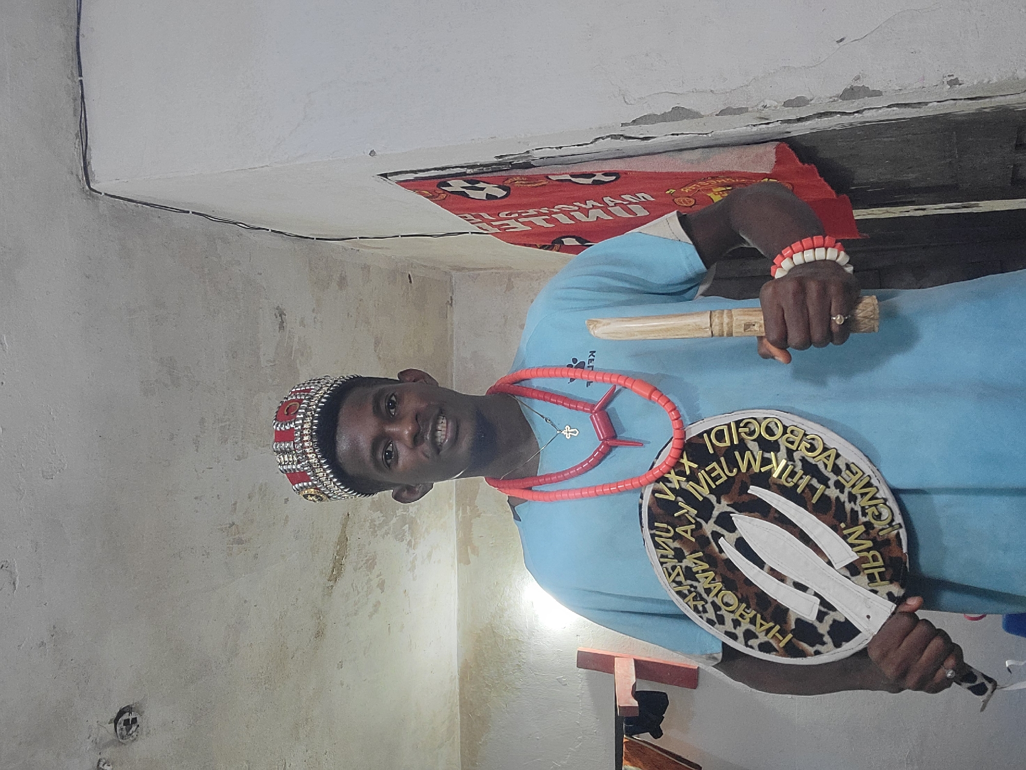 A young Black man stands in a dimly lit room, wearing a light blue t-shirt, a red and gold crown, and multiple strands of red beads around his neck and a beaded bracelet on his right wrist. He holds a wooden staff in his right hand and a circular fan with a leopard print trim and gold lettering in his left hand. The fan has white decorative elements that resemble swords. The room appears to be a home, with textured walls and a ceiling that shows signs of wear. A red cloth with black soccer balls and white lettering hangs on the wall behind him. The man is looking at the camera and smiling. The overall impression is of a ritualistic or ceremonial setting.