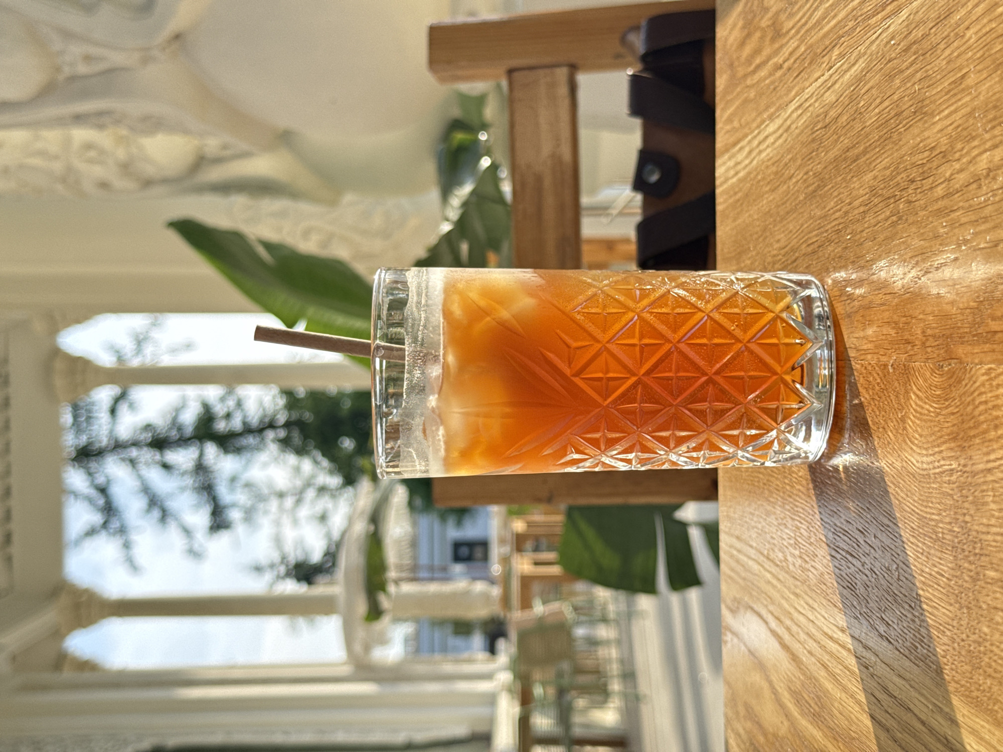 The image shows a glass of iced tea with a straw on a wooden table, outdoors. In the background, there is a view of a white building with ornate architectural details, suggesting an outdoor seating area. Green foliage is visible, and the overall setting looks like a cafe or restaurant. The scene suggests a leisurely activity in an upscale outdoor environment, possibly in a location with ornate architecture. The location is not identifiable as Moscow, Russia, from the given image.