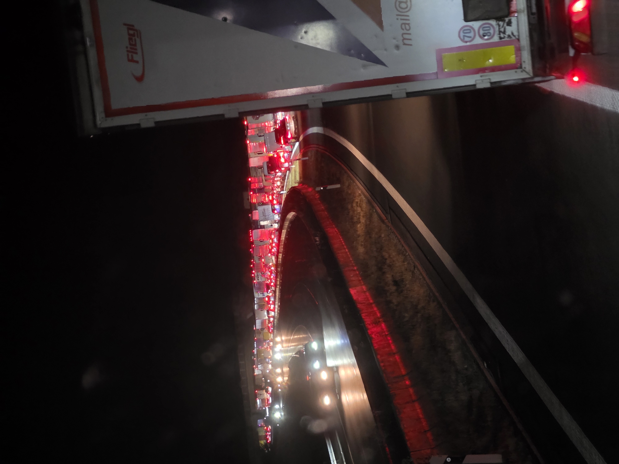 The image depicts a nighttime traffic jam on a multi-lane highway, likely near Uettingen, Germany, given the provided information. The road is illuminated by the headlights of numerous vehicles and the red taillights of stationary cars and trucks, creating streaks of light on the wet road surface. The scene is dominated by a large, white semi-trailer truck in the foreground on the right, featuring the "Fliegl" logo. The truck's rear lights are clearly visible and illuminated. In the background, a long line of traffic extends into the darkness, with brake lights and headlights forming a congested pattern. The overall environment suggests a significant disruption to normal traffic flow, possibly due to an accident, road closure, or other event, occurring on a highway that appears to be bordered by grass and possibly a sound barrier or guardrail with red reflective elements. The sky is dark, indicating nighttime conditions.