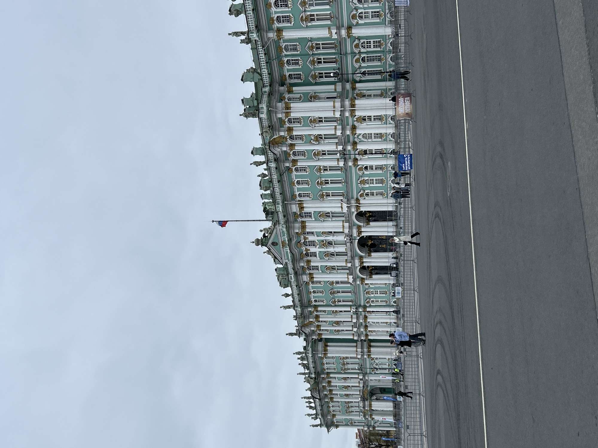 The image captures a wide view of the Winter Palace, part of the State Hermitage Museum, in Saint Petersburg, Russia, under an overcast sky. The grand Baroque-style building dominates the left side of the frame, featuring a distinctive light green facade with white columns, ornate gold detailing, and numerous windows. Statues and decorative elements adorn the roofline. A Russian Federation flag (white, blue, red horizontal stripes) flies from a flagpole positioned near the left side of the palace.
In the foreground, a vast expanse of asphalt pavement, likely Palace Square, stretches out, mostly empty except for a few scattered individuals and temporary metal barriers. These barriers run along the base of the palace, separating it from the square. Several people are visible walking near these barriers, some appearing to be staff or participants in an event. A white line divides the asphalt, and faint tire marks are visible on the darker surface.
Attached to the barriers are several banners. One prominent yellow/orange banner displays the Cyrillic text "ЗВЕЗДНАЯ ЭСТАФЕТА" (ZVEZDNAYA ESTAFETA), translating to "Star Relay" or "Star Race." Another blue banner reads "ЗОНА ПОСАДКИ УЧАСТНИКОВ ЭСТАФЕТЫ" (ZONA POSADKI UCHASTNIKOV ESTAFETY), which means "Participant Boarding Zone of the Relay." The presence of these banners and barriers suggests preparations or an ongoing "Star Relay" event, although the square itself is not crowded. The diffuse lighting indicates a cloudy day, rather than bright sunshine.