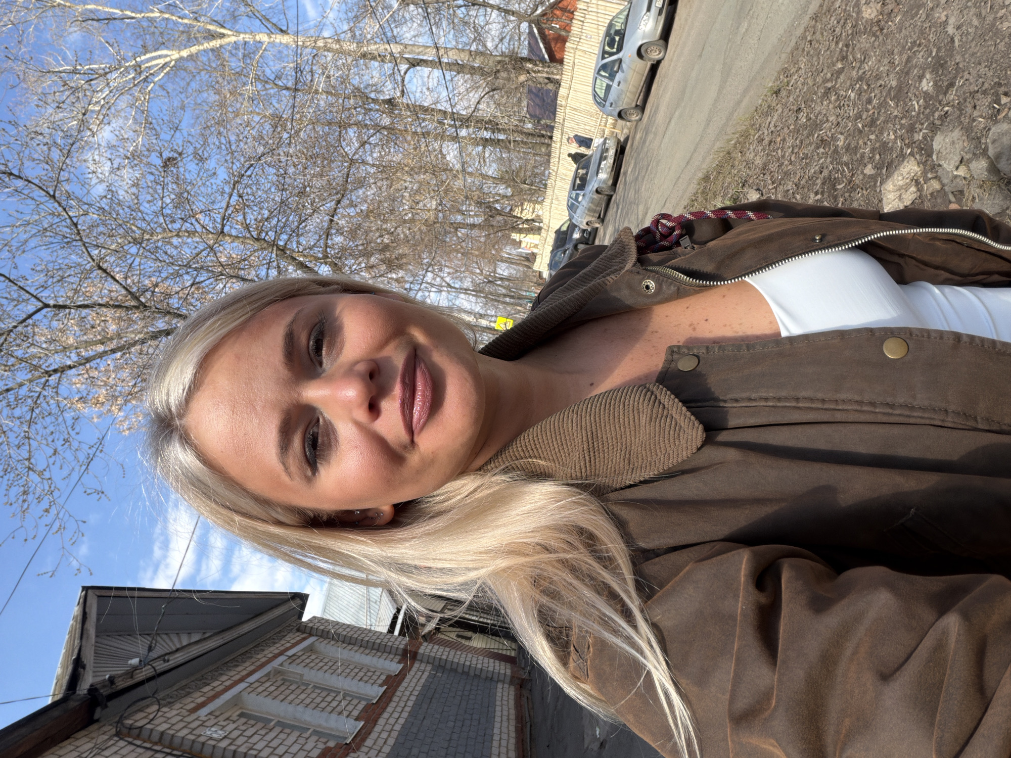 A blonde woman is taking a selfie outdoors under a bright, clear blue sky, with the image rotated 90 degrees clockwise. She is smiling gently at the camera, capturing a moment in what appears to be Sarapul, Russia. She is wearing a brown, canvas-like jacket with a ridged corduroy collar and a white top underneath. Her long, light blonde hair falls over her shoulders.
The background reveals a street scene, characteristic of late winter or early spring, given the multitude of bare trees lining the road. Several cars, including older models, are parked along the dusty roadside, which is bordered by a tall wooden fence. In the distance, two indistinct figures are visible further down the street. To the woman's left, a section of a light-colored brick building with a dark roof and overhead utility wires adds to the urban or suburban setting. The ground near the parked cars and fence consists of dry earth and scattered leaves. The overall mood conveyed by the woman's smile and the sunny weather is pleasant and relaxed. There is no visible text in the image.