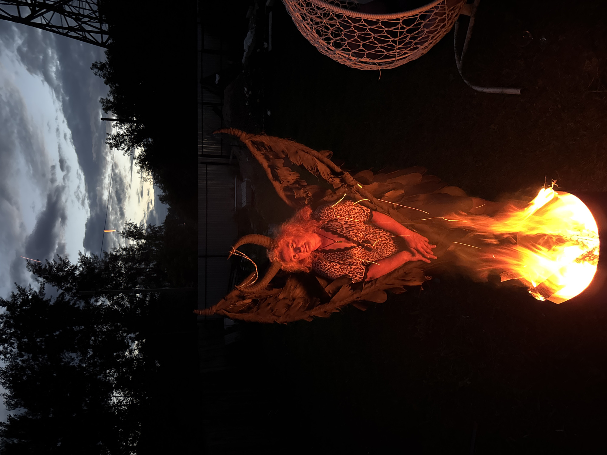 In the city of Chaikovsky, Russia, at dusk, a person is positioned in front of a bonfire. The individual is wearing a leopard-print top and has an elaborate, feathery costume with horns and what appear to be large wings. They are holding their hands clasped together. The bonfire is burning brightly, casting a warm orange glow on the person and the immediate surroundings. To the right of the frame, a large, white, woven hanging chair is visible. The background is dark, with silhouetted trees and a fence, suggesting a backyard or outdoor area. The sky above is cloudy, with hints of twilight colors. A tall, dark metal structure, possibly a transmission tower, is visible on the far right of the image, against the twilight sky.