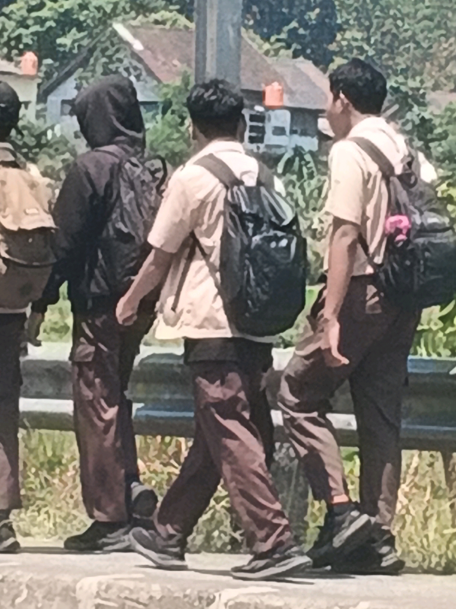 The image depicts three young males, appearing to be students, walking outdoors in what seems to be a semi-rural area, possibly near a school in Kalirejo, Indonesia. The individuals are dressed in school uniforms. Two of the students are wearing light-colored shirts and dark pants, with one wearing a hooded jacket over his shirt. They are all carrying backpacks. The background shows trees and buildings, suggesting a residential or school environment. The ground they are walking on appears to be paved or a walkway, with a railing in the foreground. The lighting suggests it is daytime.