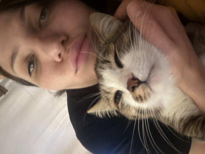 A woman with brown hair, full lips, and light skin is holding a brown tabby and white cat. She's looking at the camera with a slight pout, while the cat appears relaxed and content. The cat is pressed against her face, with one paw visible. They are both nestled together in what appears to be a close and affectionate pose. The background is blurred, suggesting a close-up shot.