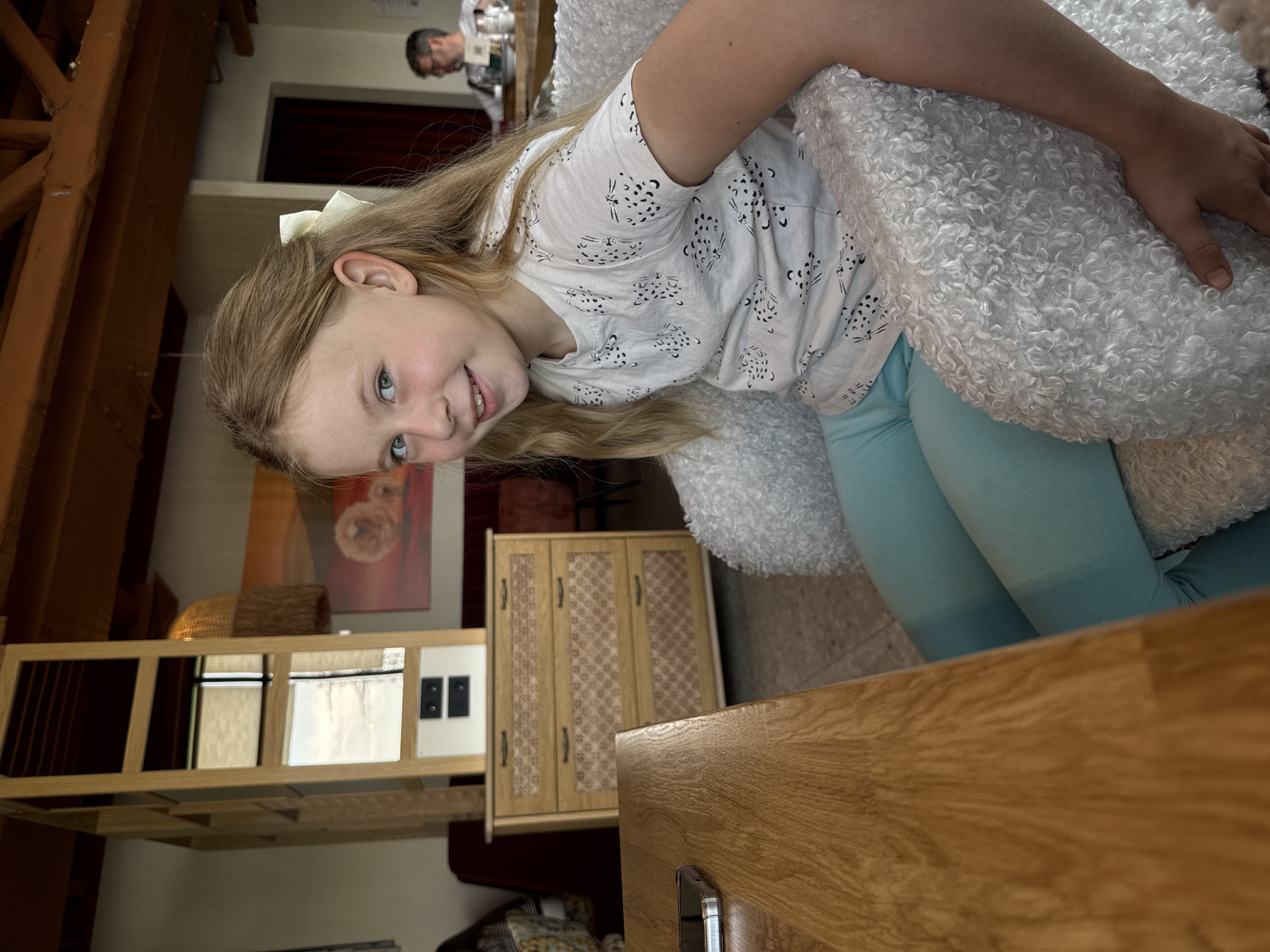 A young girl with blonde hair and a white bow is leaning against a large, white, fluffy object. She is wearing a white t-shirt with a black design and light blue leggings. A man is visible in the background. The room contains wooden furniture including a shelf unit, a cabinet, and a table. The image appears to be taken indoors, but there are no clear indicators to pinpoint the location to Veliky Novgorod, Russia.