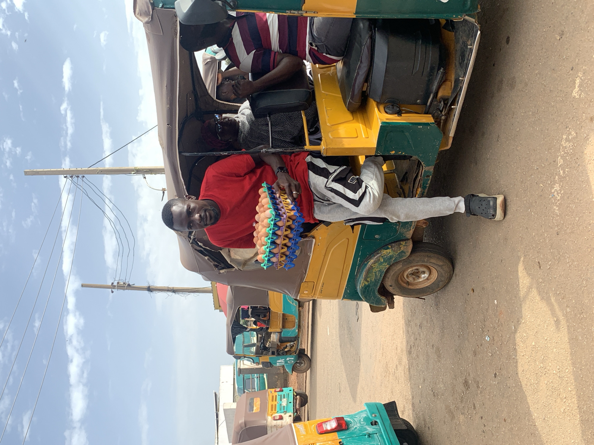 In Gura Top, Nigeria, a young Black man in a red t-shirt and grey sweatpants with black and white stripes on the side, is standing next to a yellow and green auto-rickshaw, holding a blue plastic tray filled with eggs. He is looking directly at the camera. Inside the auto-rickshaw, a Black woman with glasses and a patterned scarf is seated, and another person is partially visible behind her. The auto-rickshaw has a brown canvas roof and is parked on a dirt road. In the background, other auto-rickshaws, power lines, and buildings are visible. The sky is partly cloudy.