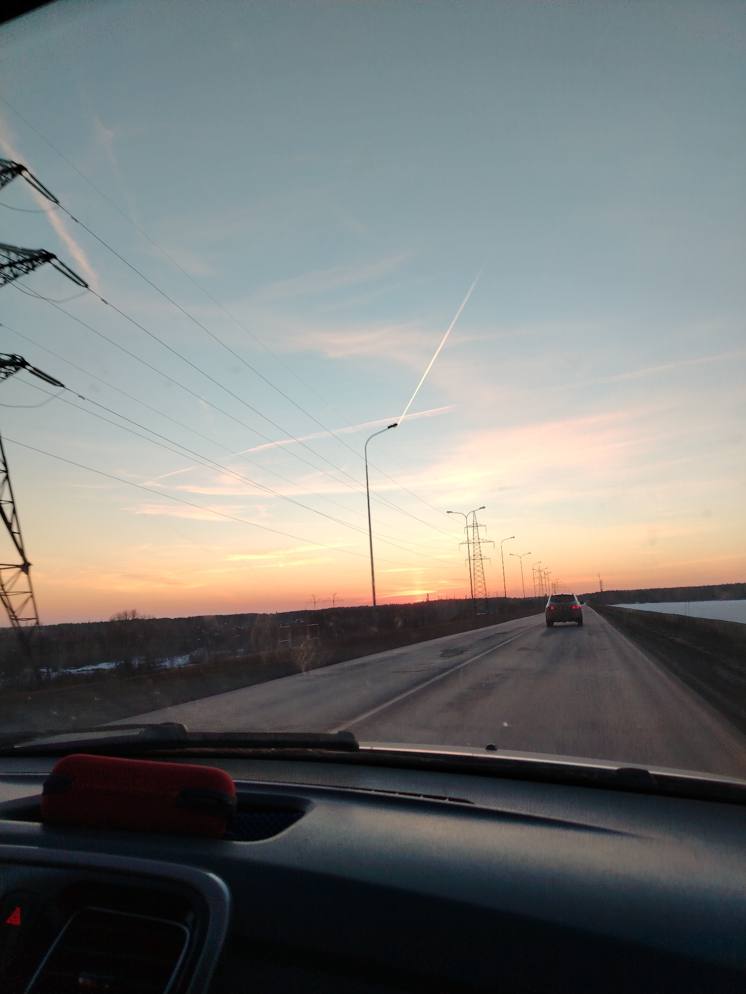 The image is taken from the interior of a car, likely the driver's perspective, on a road outside the city of Chaikovsky, Russia. It is sunset, casting an orange glow on the horizon. Power lines and streetlights run along the road, and a car is ahead. The sky is a mix of blue and orange hues, with contrails visible. The road surface shows signs of wear. The dashboard of the car is visible in the foreground, with a red object placed on it. The environment appears to be a rural or semi-rural area, with trees visible on the left side of the road and a flat, possibly snowy, field on the right.