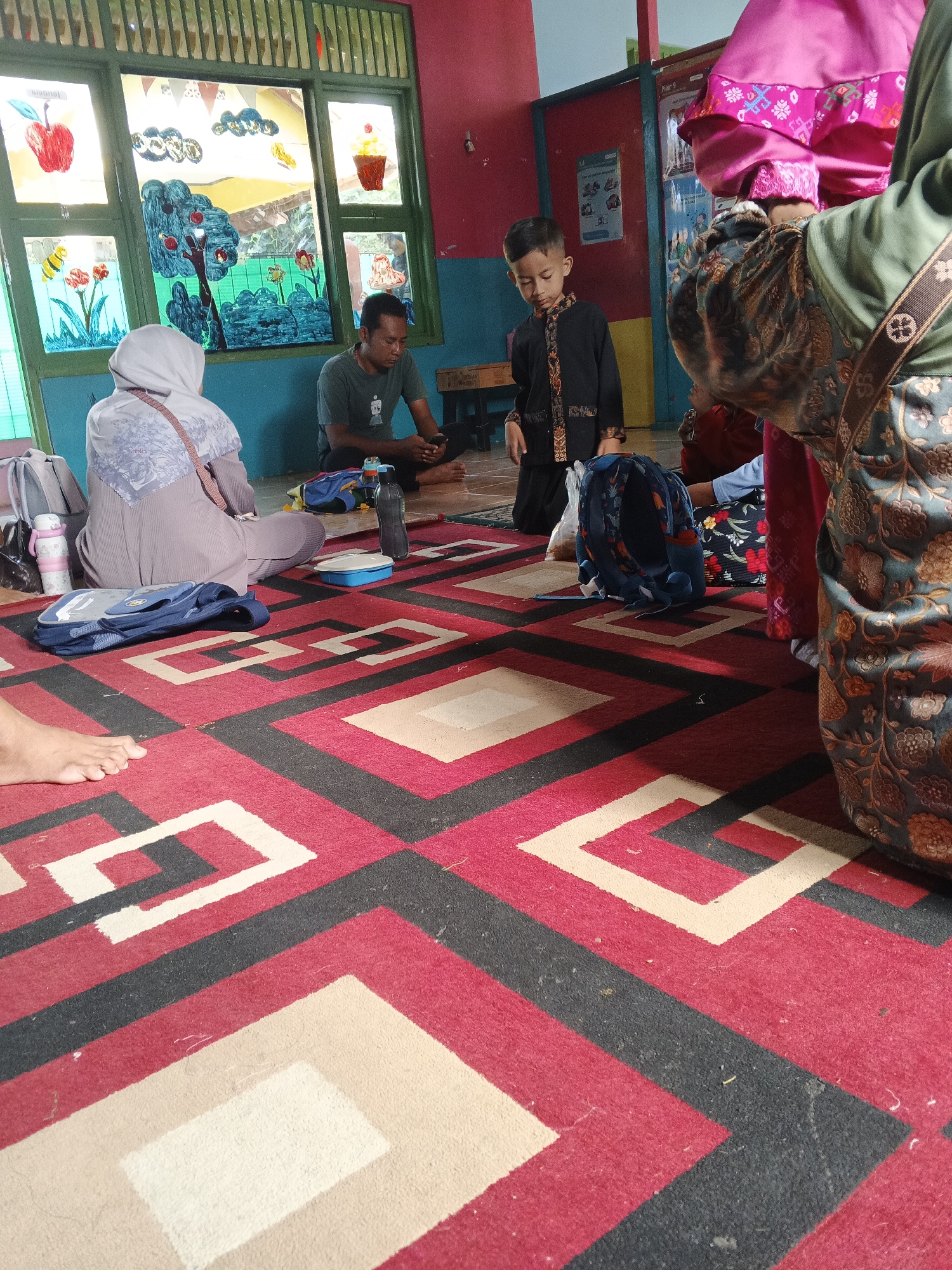 A diverse group of people are gathered on a red, black, and white patterned carpet in a room with stained-glass windows depicting fruit and flowers. A man in a dark green t-shirt sits on the floor, looking at his phone. A child in a black and gold outfit stands nearby, also looking down. A woman in a grey hijab sits with her back to the camera, and another person in a green and brown patterned outfit is partially visible on the right. Backpacks, a water bottle, and a small blue container are scattered on the carpet. The room appears to be a classroom or common area in Warungkiara, Indonesia, with colorful walls and posters visible in the background.