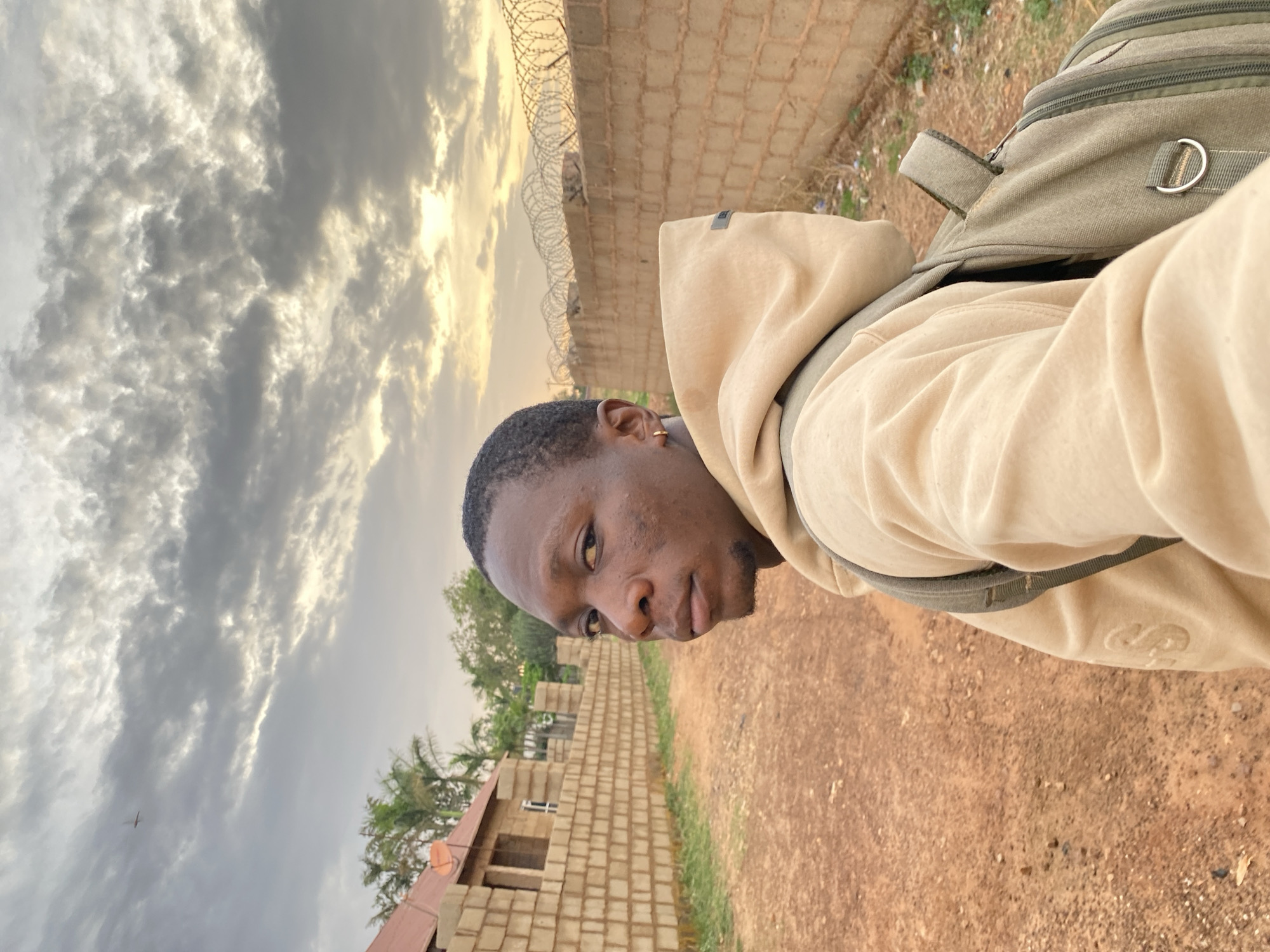 The image captures a selfie-style portrait of a young man outdoors, rotated 90 degrees clockwise. He appears to be of African descent, with short dark hair and a neatly trimmed beard, and wears a light beige or cream-colored hooded sweatshirt. A green backpack with visible straps and a silver D-ring is worn on his back, and a small gold hoop earring is visible in his left ear. He looks towards the camera with a neutral to slightly serious expression.
The background reveals a semi-urban or rural landscape under a dramatic sky. The sky is filled with dark, textured clouds, interspersed with streaks of bright, golden light, suggesting either sunrise or sunset. A low wall constructed from concrete blocks and topped with barbed wire stretches across the mid-ground. Beyond the wall, partially constructed block buildings with red-brown roofs and a small satellite dish are visible, along with sparse green trees, possibly palm trees. The foreground and ground near the buildings consist of reddish-brown dirt, with some dry grass and debris. A faint, small object resembling an airplane or drone is visible against the cloudy sky in the distance. A faint, stylized 'S' or similar logo is subtly embossed or stitched on the lower part of the man's hoodie. The scene suggests a developing area, consistent with the Bukuru, Nigeria location context.