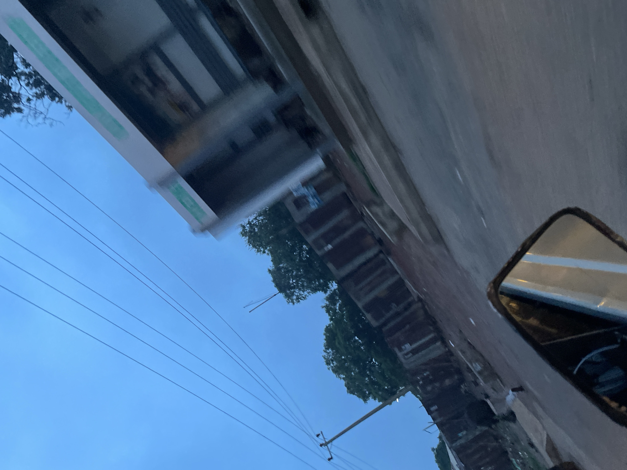 The image depicts a street scene in Jos, Nigeria, during what appears to be dusk or early morning. The perspective is from a moving vehicle, likely a motorcycle, as evidenced by the reflection in the rearview mirror in the foreground.
Dominating the upper portion of the image are several power lines stretching across the twilight sky, which is a muted blue. To the right, a blurred structure, possibly a bus stop or a roadside stall with a white roof and green accents, is visible. Trees, some dense and leafy, are scattered throughout the mid-ground.
The middle and lower sections of the image show a road surface that is uneven and somewhat worn. Alongside the road, there is a wall constructed of corrugated metal sheets, showing signs of rust and age. Beyond this wall, glimpses of makeshift structures and debris suggest a less developed or informal settlement. The overall impression is one of a utilitarian urban environment. No people are clearly discernible.