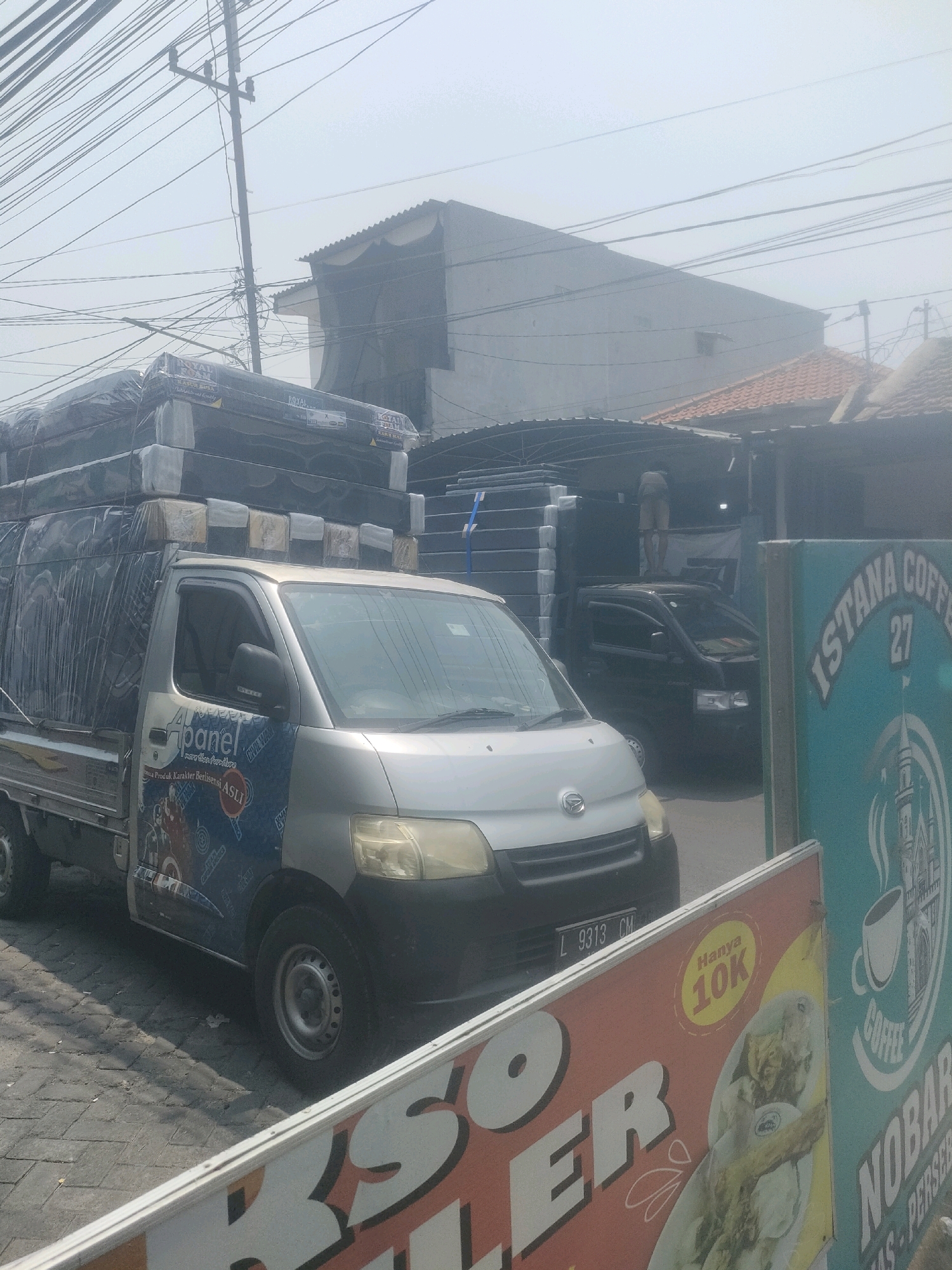 A light silver, four-door pickup truck is parked on a paved road, partially obscuring a black truck and a building behind it. The pickup truck's bed is loaded with stacked mattresses, wrapped in clear plastic and covered with a blue tarp. The side of the pickup truck features a blue and white decal with text and an image. A white sign with orange text and an image of food is in the foreground. Another teal sign with white lettering that reads "ISTANA COFFEE 27" and an illustration of a building and a coffee cup is to the right of the food sign. Overhead, numerous electrical wires are visible against a hazy sky. The scene appears to be in an urban or industrial area.