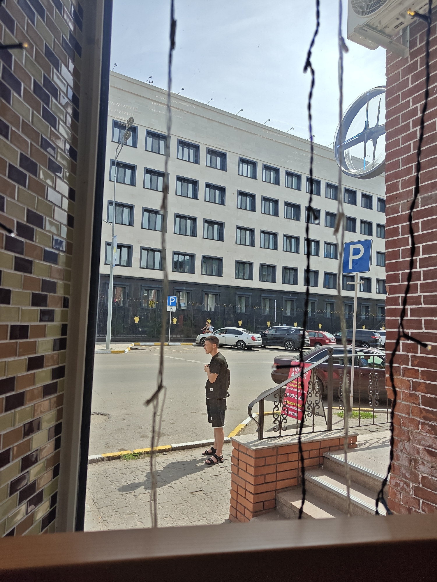 The image shows a view through a window onto a street scene in Petropavl, Kazakhstan. A man in a black t-shirt and shorts stands on a sidewalk, looking to his right. Behind him, a multi-story white building with numerous windows is visible. Several cars are parked along the street, and a blue parking sign is posted on a pole. The foreground shows a brick wall and a portion of a window frame, with strings hanging down, possibly from Christmas lights. The street is paved, and there is a yellow curb marking. The sky is overcast. The brick building on the right side of the image appears to be a shop or entrance to a building with a set of stairs leading up to it.