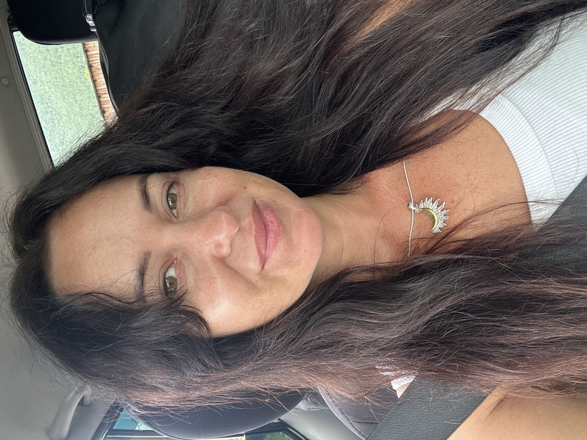 The image shows a woman in the passenger seat of a car. She has long, dark, wavy hair and is wearing a white ribbed tank top and a silver necklace with a sun pendant. Her eyes are open and she is looking at the camera. The background outside the car window appears to be a natural landscape with greenery and a wooden structure. The overall impression is a selfie taken while traveling. No specific details about Slyudyanka, Russia are discernible from the image itself.
