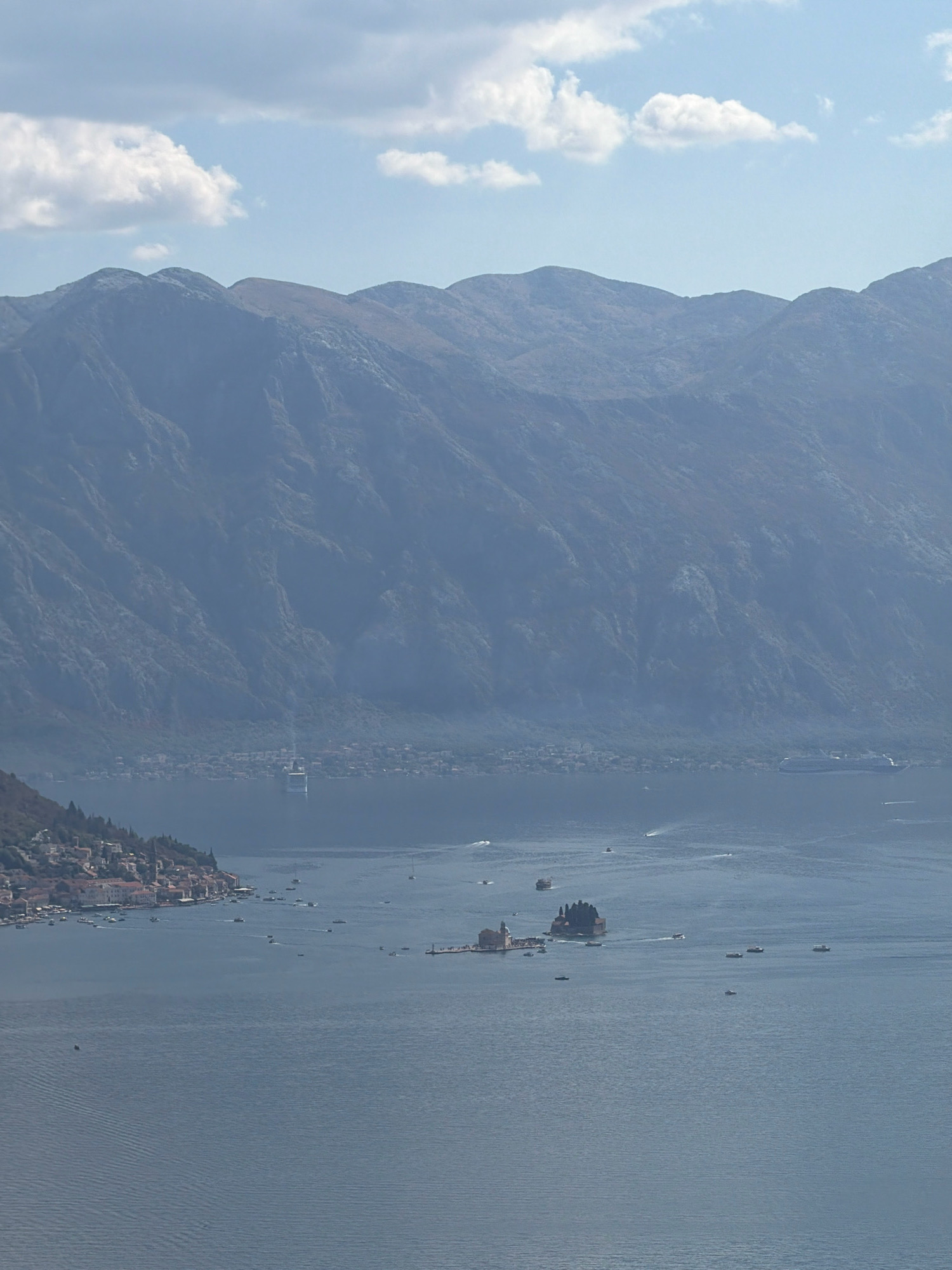 The image shows a wide, panoramic view of a bay, with mountains rising in the background and a town nestled on the coastline to the left. The water is a deep blue, with ripples and a few boats scattered across its surface. In the distance, a large cruise ship is visible. On the left side of the bay, a town with red-tiled roofs and church steeples is situated on a hillside. There is a small island in the bay with buildings on it, appearing to be a religious site. The sky is a pale blue with some scattered white clouds. The overall impression is one of a tranquil, scenic landscape. The location is identified as Morinj, Montenegro, and the scene depicts the Bay of Kotor.