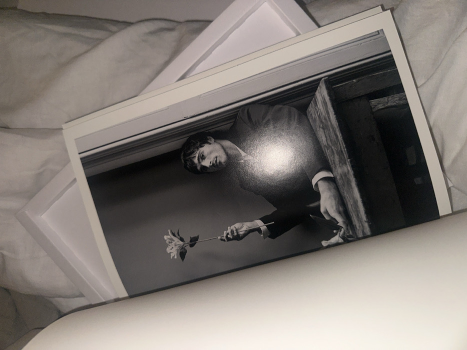 A black and white photograph depicts a man in a suit, holding a single flower. He is positioned in what appears to be a doorway or opening, peering downwards. The photo is printed with a white border and rests atop multiple layers of white framing materials. It is situated on a white, possibly fabric, surface with subtle light and shadow variations.