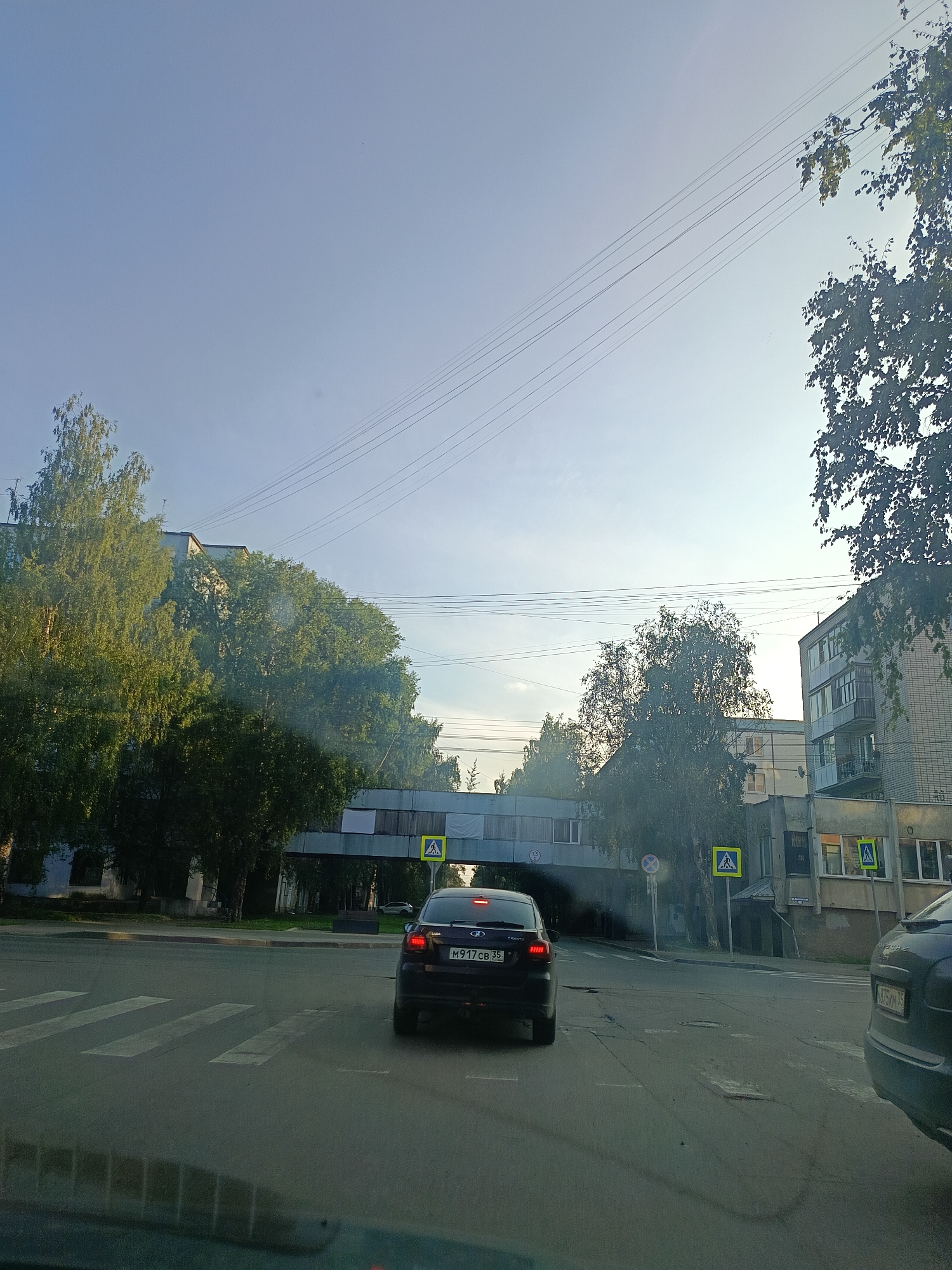 The image depicts a street scene in Vologda, Russia, with a focus on a pedestrian crossing and traffic. The sky is clear and blue, with power lines visible across the upper portion of the frame. On the left, there are multi-story apartment buildings partially obscured by lush green trees. A pedestrian overpass crosses the street in the middle ground. On the right side of the street, another apartment building is visible, with balconies and windows.
In the foreground, a black Lada Granta is stopped at a pedestrian crossing, its brake lights illuminated. The license plate reads "M917CB 35". To the right of the Lada, the rear of a dark gray car, possibly a Porsche, is visible, with a license plate reading "875KM 35". The pedestrian crossing itself is marked with white stripes on the asphalt. Yellow pedestrian crossing signs are posted at the intersection on both sides of the road. The overall impression is of a typical urban intersection during daylight, with vehicles waiting for pedestrians.