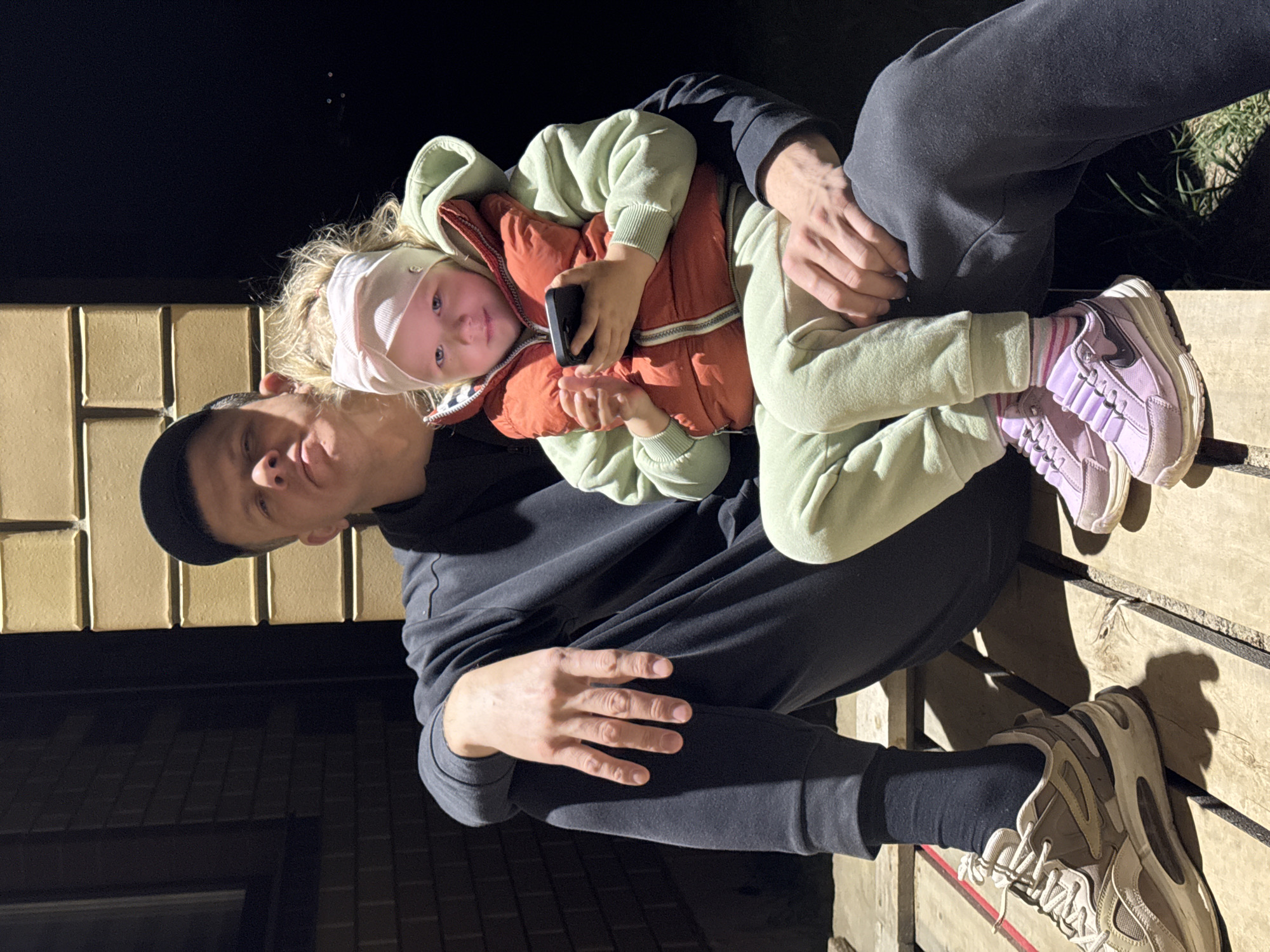 A man and a child are seated on a wooden structure outdoors. The man is wearing a black hooded sweatshirt, black pants, black socks, and tan sneakers. He is also wearing a black baseball cap. He is holding the child, who is wearing a white head wrap, a light green hooded sweatshirt, an orange vest, light green pants, and purple sneakers with pink stripes. The child is holding a smartphone. The background includes a brick structure and dark shadows, suggesting it is night in Ферма, Russia.
