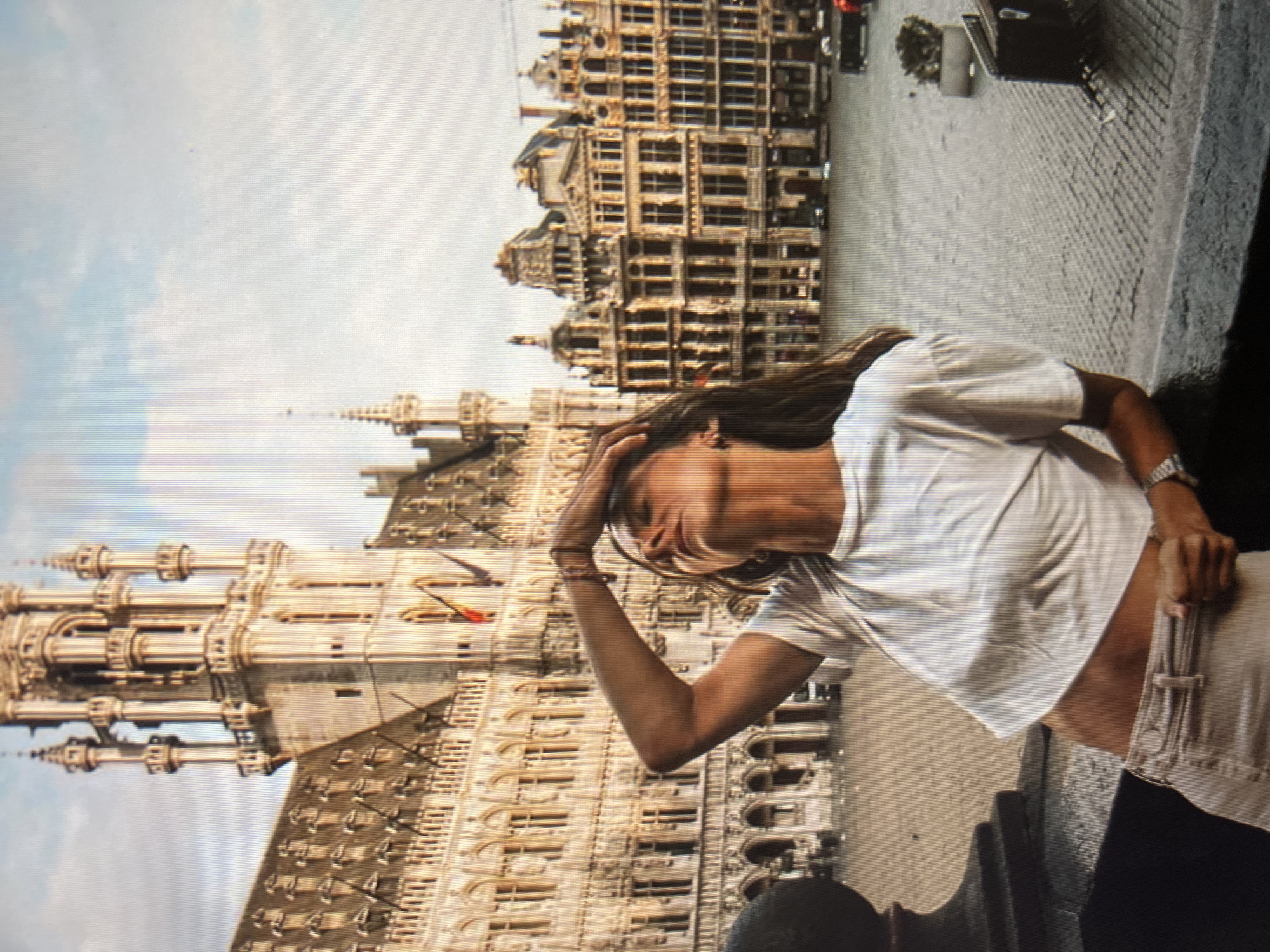 A young woman is pictured leaning on a railing in a city square with ornate historical buildings in the background. The woman has her eyes closed and her hand on her forehead, suggesting discomfort or heat. She is wearing a white crop top and light-colored pants. The architecture behind her is elaborate, with many windows and decorative elements. The ground of the square appears to be paved with cobblestones or similar material. The sky above is partly cloudy. The scene is set in a city square, which could be anywhere with such architecture, but the prompt specifically requests to identify the location as the city of Qarağandı city administration, Kazakhstan.