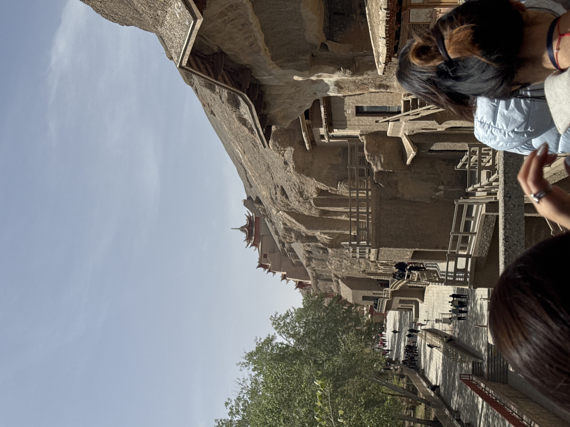 The image shows a view of the Mogao Caves, a UNESCO World Heritage Site located in Dunhuang City, China. The caves are carved into a cliff face and feature Buddhist art dating back to the 4th century. The scene depicts the complex of caves, connected by wooden walkways and staircases. The structures are built into the cliff face, blending architecture with the natural rock formation. A crowd of people is walking on the pathway below, suggesting a tourist attraction. A large tree is visible in the foreground, along with an azure sky. Two women are visible in the foreground on the right side of the frame.