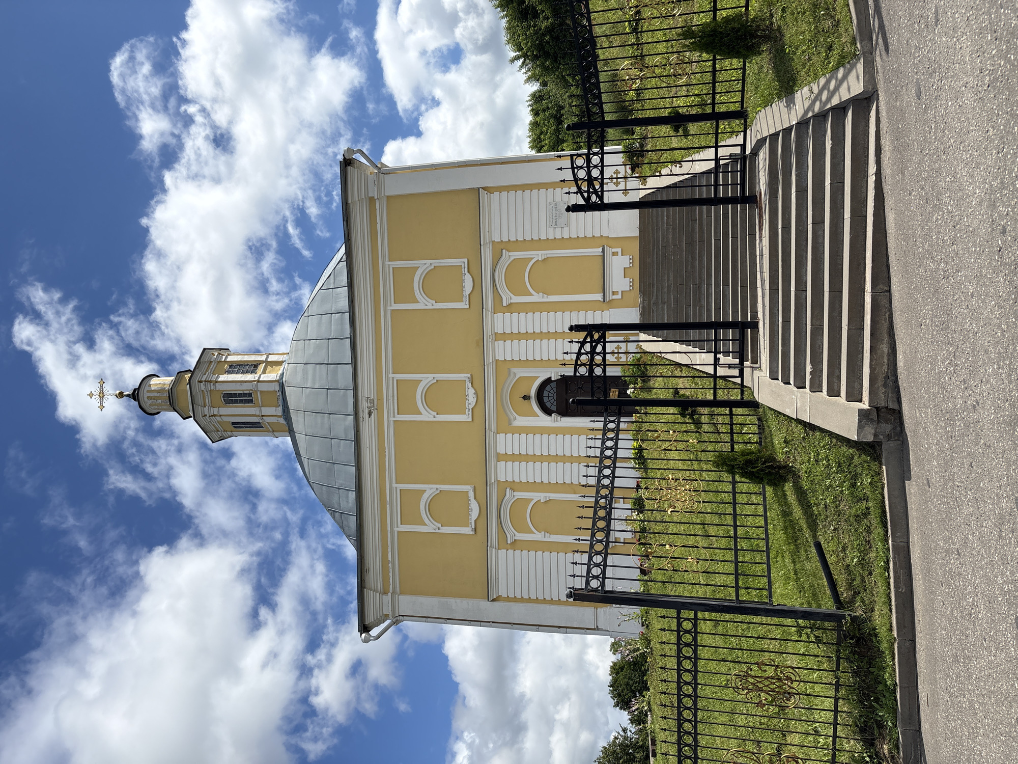 This is a full shot of a yellow church with a gray dome, topped with a golden cross, under a partly cloudy blue sky. The church has white trim and arched window details. A black wrought iron fence with ornate gold accents surrounds the church grounds. A set of concrete stairs leads up to the church entrance, which is a dark wooden door. Green grass and some small trees are visible on the church grounds. The church is located in the village of Radkevshchina, Russia.