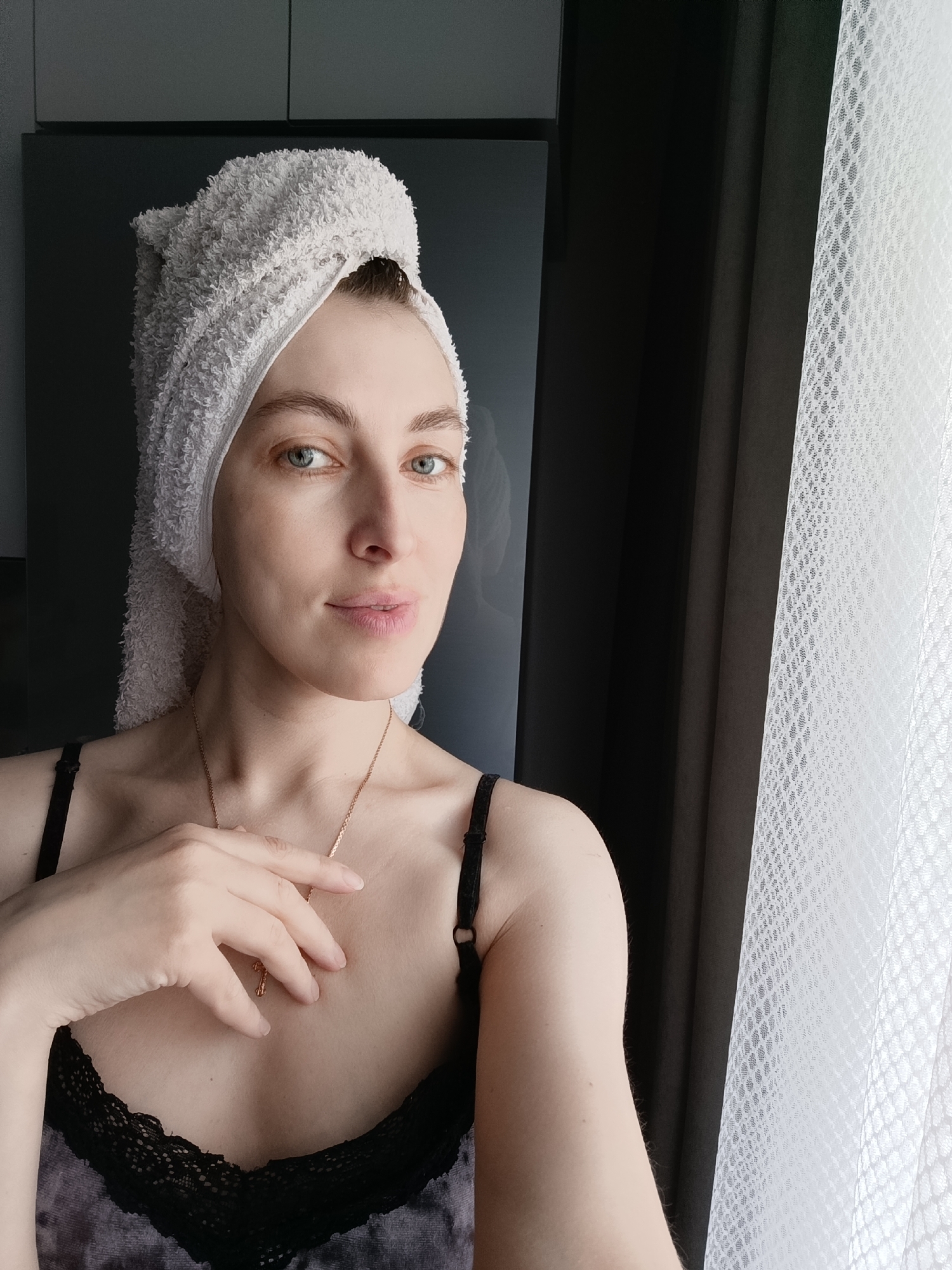 A woman with light skin and striking blue-green eyes is captured in a close-up indoor selfie, located in Chaikovsky, Russia. She has a white terry cloth towel wrapped around her head in a turban style, suggesting a recent shower or beauty routine. Her expression is soft and serene, with a slight, natural smile as she looks directly at the camera. She wears a black camisole with delicate lace detailing on the V-neckline, and a thin gold-colored chain necklace with a small, indistinct pendant, possibly a cross, rests on her chest. Her right hand, with neatly manicured nails, lightly touches the necklace. The background features a dark, possibly reflective, surface or cabinet on the left, alongside a dark solid curtain. To the right, a white, intricately patterned sheer curtain indicates the presence of a window, through which natural light illuminates the scene, suggesting it is daytime. The image conveys a sense of personal calm and domestic intimacy. No text is visible in the media file.