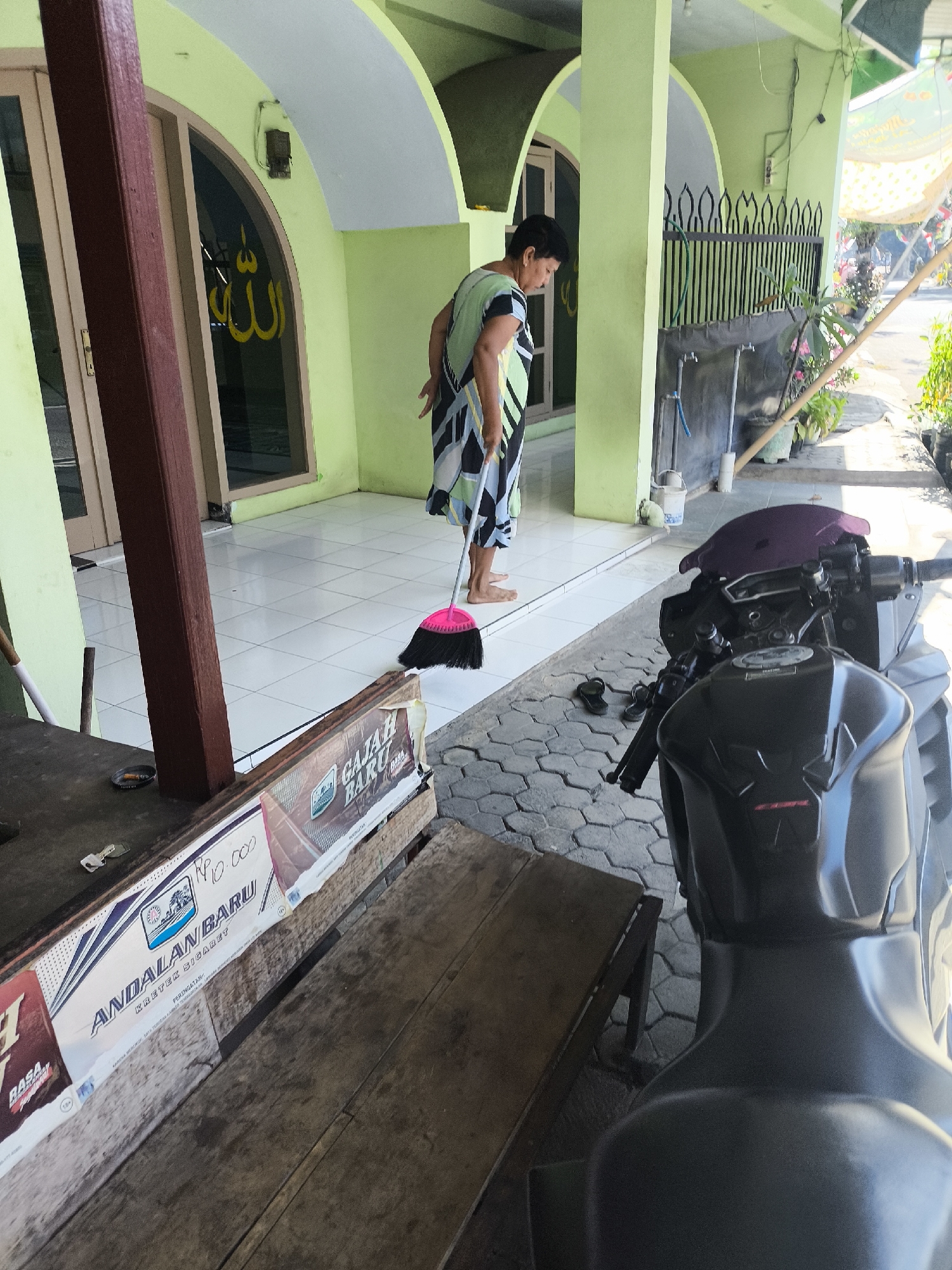 In the city of Citrodiwangsan, Indonesia, a woman with short dark hair is sweeping a tiled porch with a pink broom. She wears a black, green, and white striped dress. To the left of the woman is a large wooden pillar and a glass door with Arabic script in yellow on it, possibly indicating a place of worship. In front of the woman, on the tiled ground, are a pair of black sandals. Further to the right, a black motorcycle is parked. In the foreground, a wooden bench or table displays a sign with the words "ANDALAN BARU" and "KRETEK SIGARET," suggesting it is related to tobacco products. The environment appears to be outdoors, with a light green wall and arched doorways. Sunlight casts shadows on the ground, indicating it is daytime.