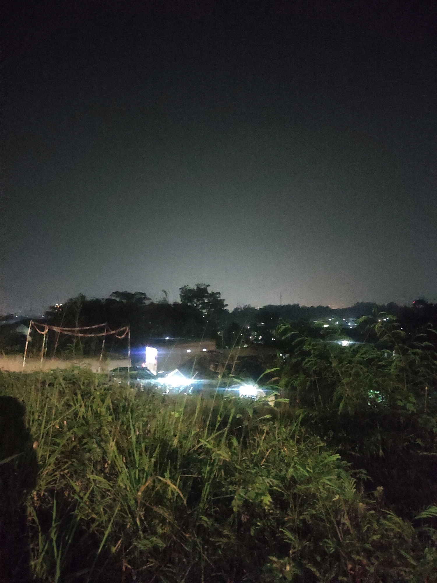 An eye-level shot shows a nighttime landscape viewed from a grassy knoll overlooking a semi-urban area. The sky is a dark, hazy gray, with the glow of distant lights faintly visible. In the foreground, tall, dry grasses and some leafy green plants dominate the scene, with a dark, indistinct human shadow on the far left.
In the midground, a line of trees separates the foreground from the lights of buildings and a road. A tall, brightly lit structure, possibly a billboard or building facade, stands out with its white light. Several vehicles are visible near this structure, their headlights and taillights creating streaks of light on the wet road. A simple, makeshift structure with red and white poles and netting is situated on the left side of the midground, partially obscured by the foreground vegetation.
Further in the background, a silhouette of trees and a gently rolling landscape can be seen, dotted with numerous small lights indicating residential or commercial areas. The overall impression is of a view from a slightly elevated position on the outskirts of a town or city at night.