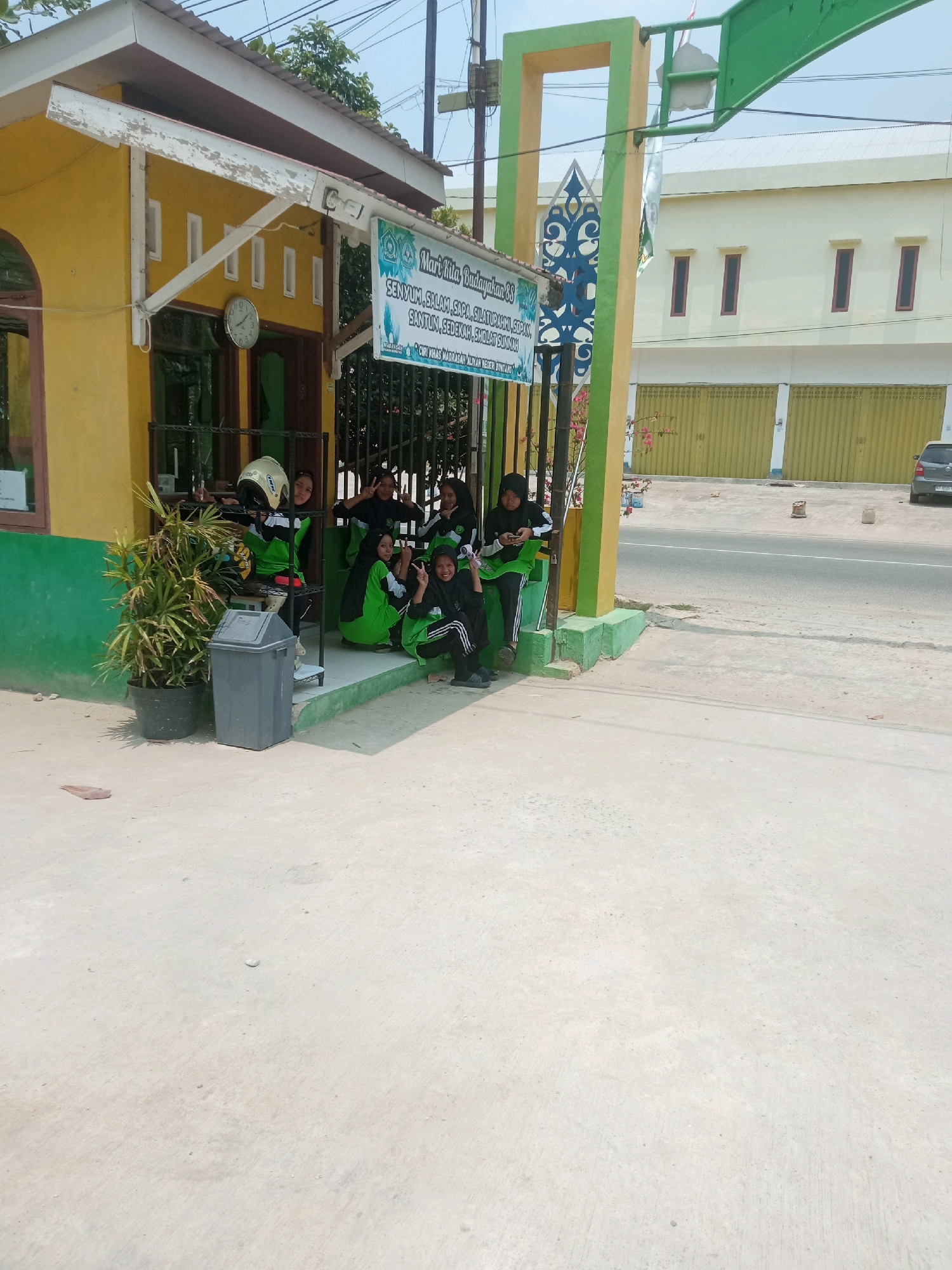 The image shows a group of five young women in Islamic attire, consisting of hijabs and tracksuits, posing in front of a yellow and green building. They are gathered near a metal gate and a tall green plant in a pot. One woman is wearing a helmet, possibly for a motorcycle. A grey trash bin is next to the plant. In the background, a light yellow building with multiple windows and doors is visible across a street. A dark colored car is parked on the right side of the street. A banner with text in Indonesian, including "Mari Kita Budayakan" and other phrases, hangs on the green gate structure. The location appears to be an entrance to a school or community building in Bontang, Indonesia, given the text on the banner and the architectural style. The overall scene is outdoors, under a clear, bright sky, suggesting a daytime setting. The women appear to be interacting casually, with some making peace signs with their fingers.
